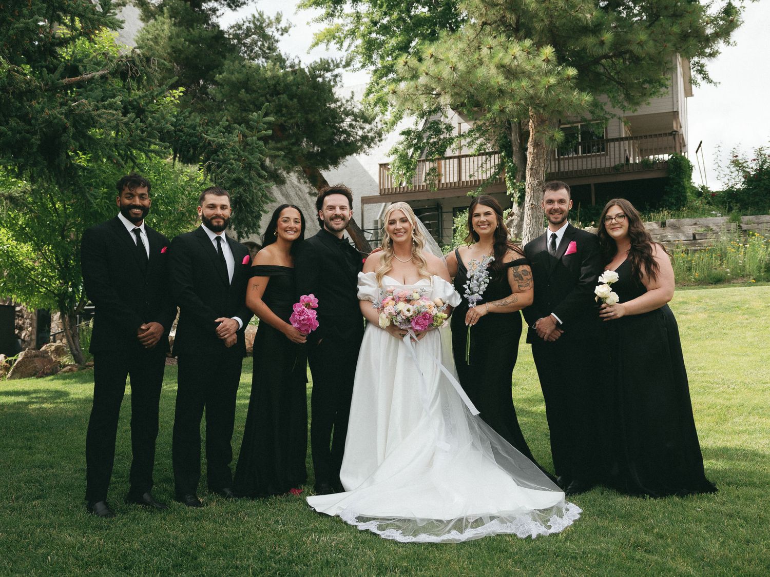 Wedding party in black formal attire with bride in white gown posing outdoors in lush garden setting.