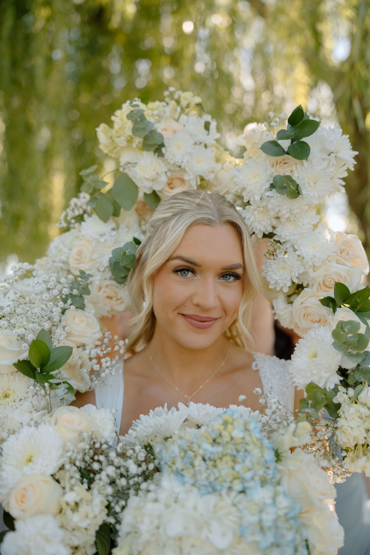 Blonde bride smiling through circular white floral arch with roses and hydrangeas holding bouquet.