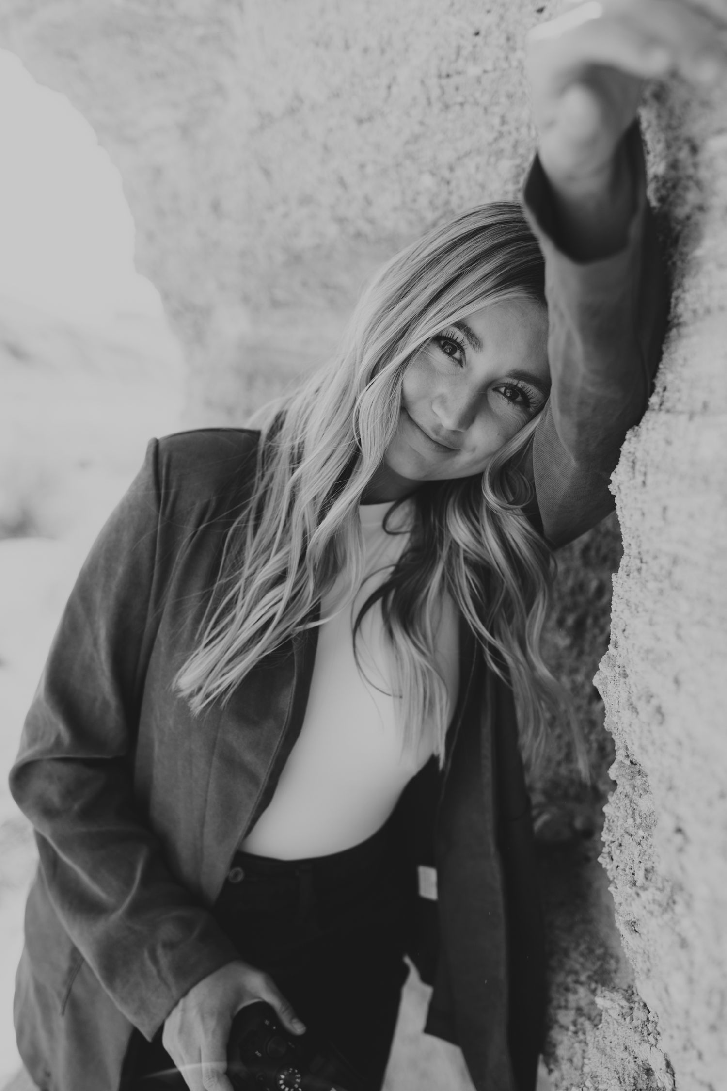 Black and white portrait of a blonde woman leaning against a stone wall in a blazer.