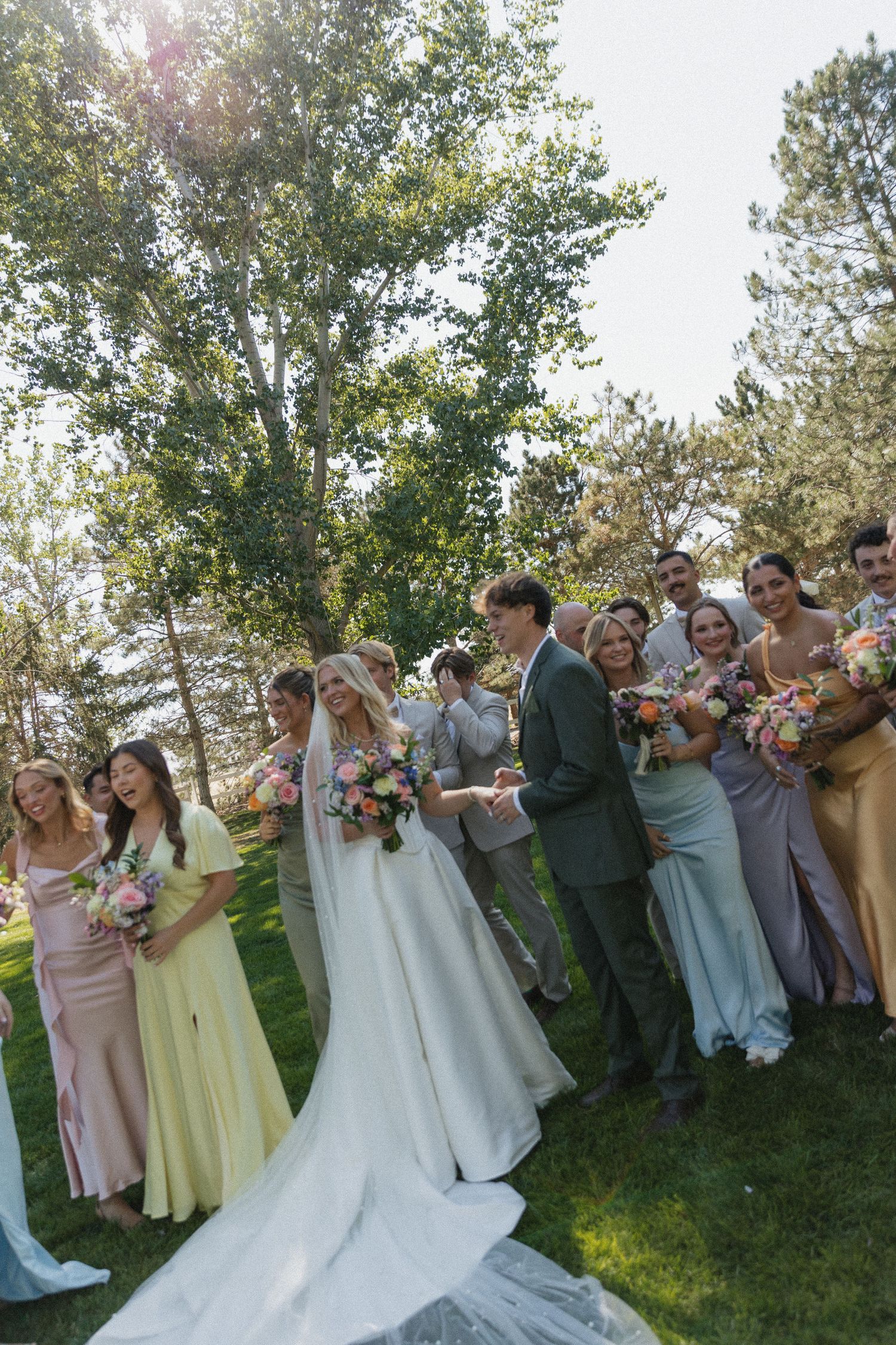 Wedding party in pastel gowns and suits celebrating with bride and groom outdoors among green trees.