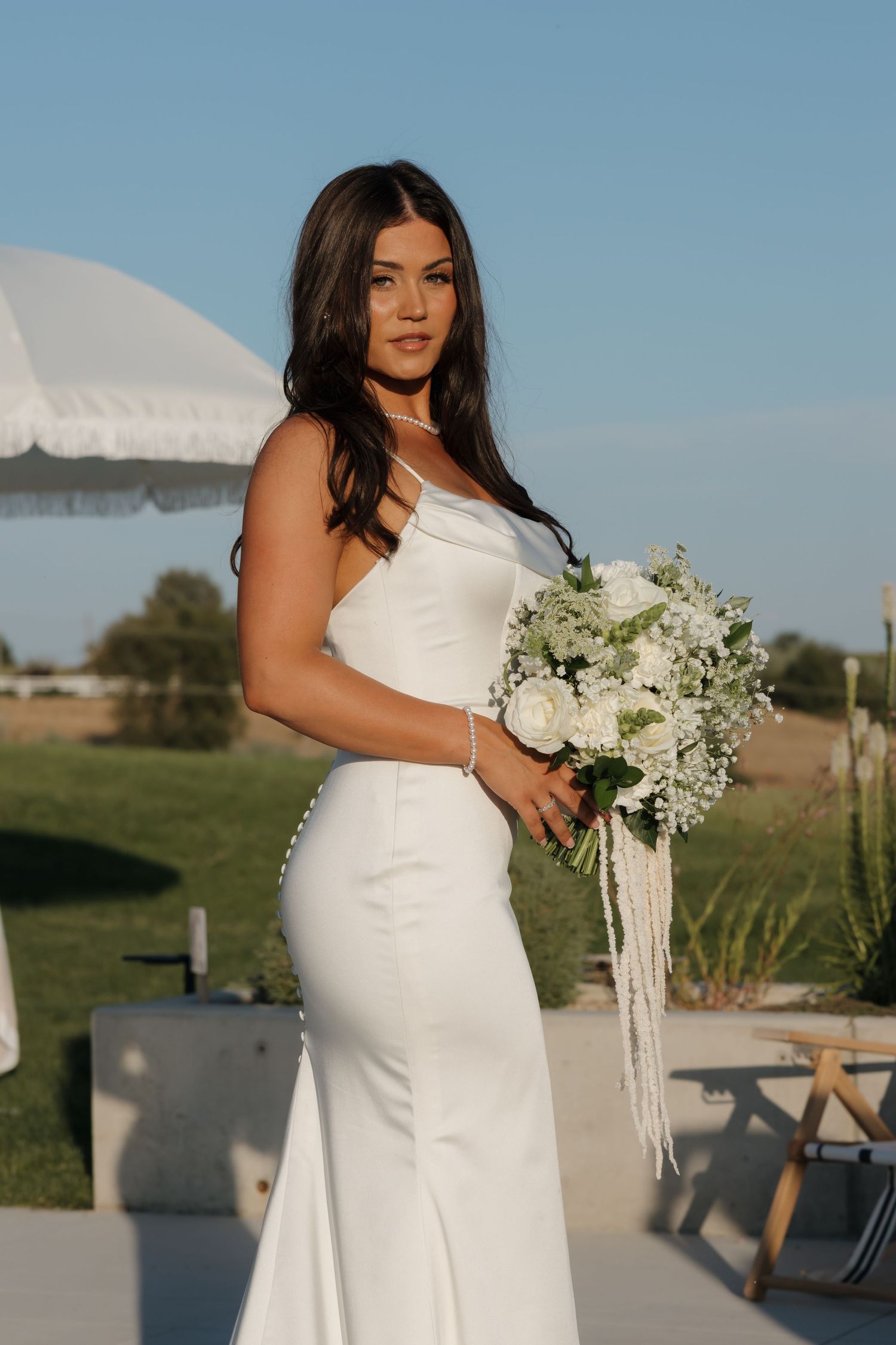 Bride in sleek white mermaid gown holding white floral bouquet outdoors at golden hour.