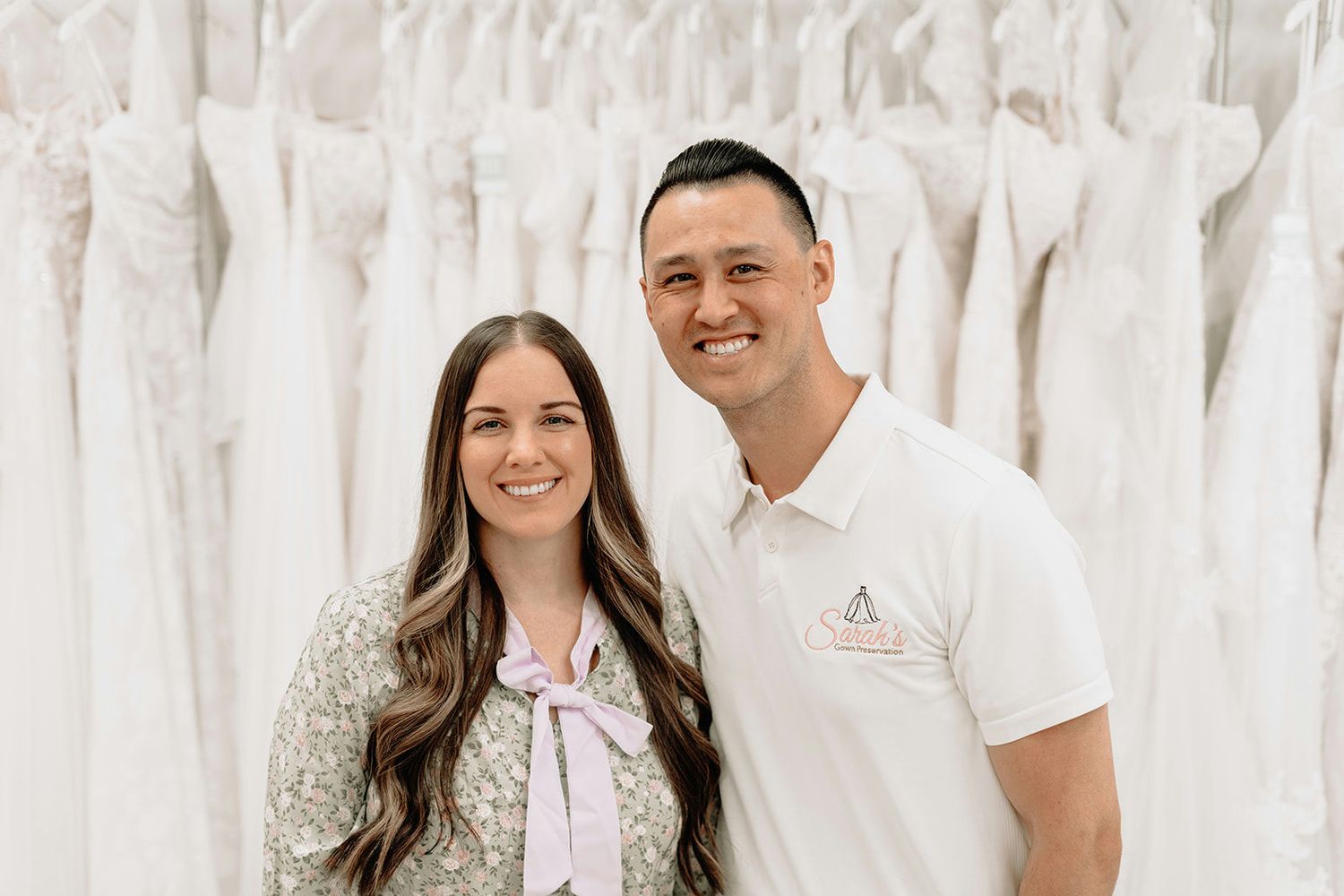 A smiling couple poses in a bridal boutique surrounded by white wedding gowns on display racks.