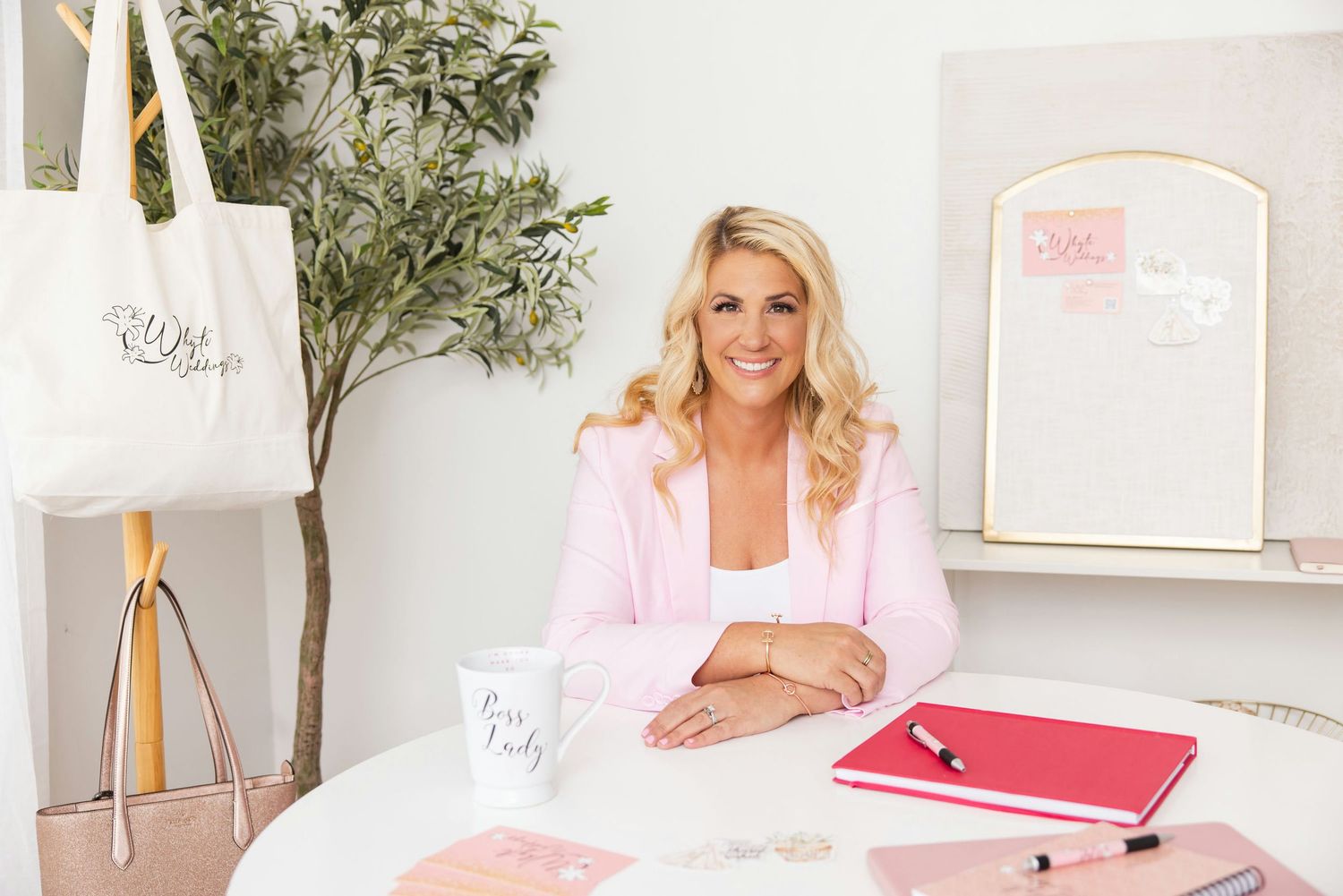 Blonde woman in pink blazer sits at white desk with planner, mug, and tote bag in bright office.