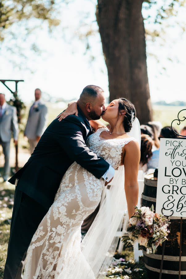 Bride and groom sharing first kiss at outdoor wedding ceremony near rustic sign.