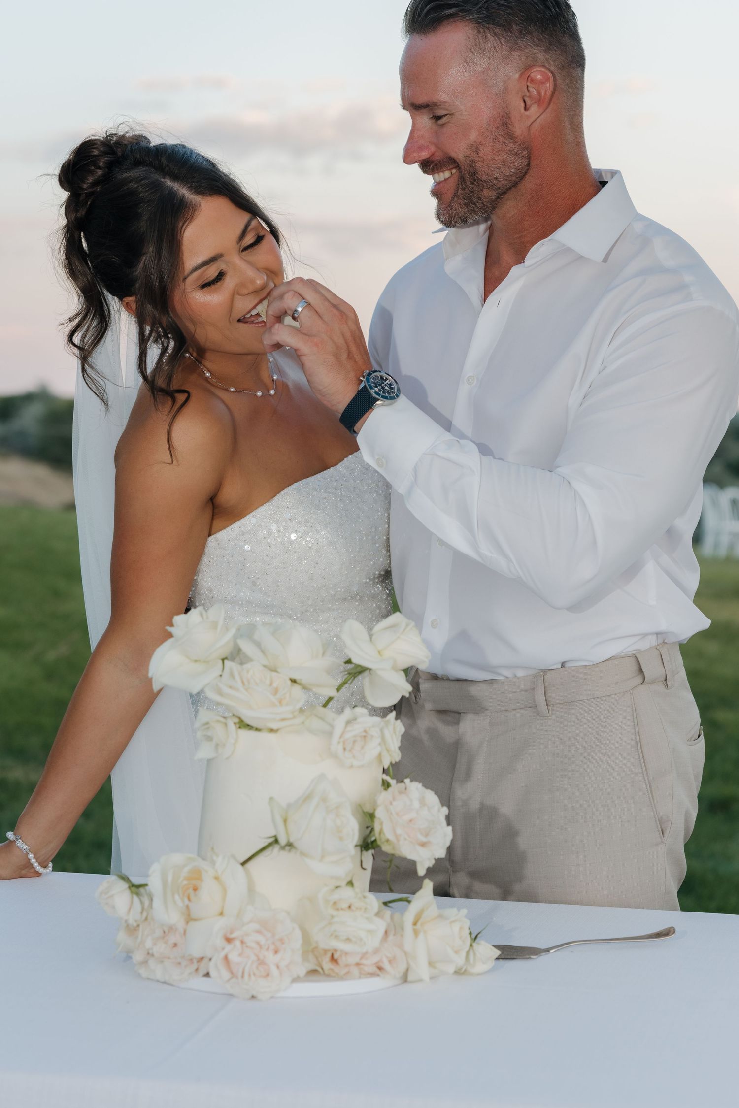 Groom feeding wedding cake to smiling bride beside elegant white rose-decorated tiered cake at sunset.