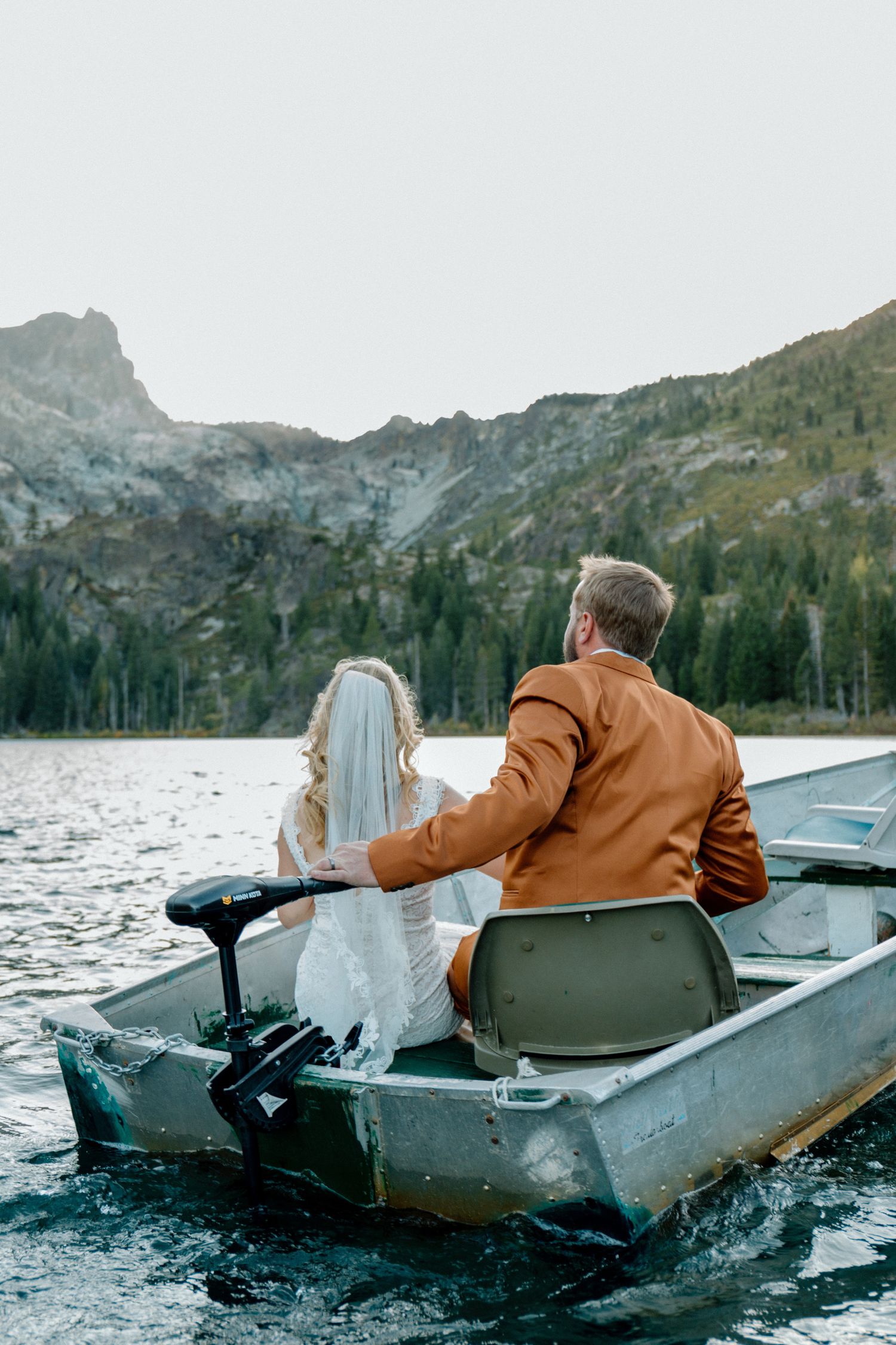 Bride and groom riding a small fishing boat on a mountain lake surrounded by pine trees and rocky peaks.