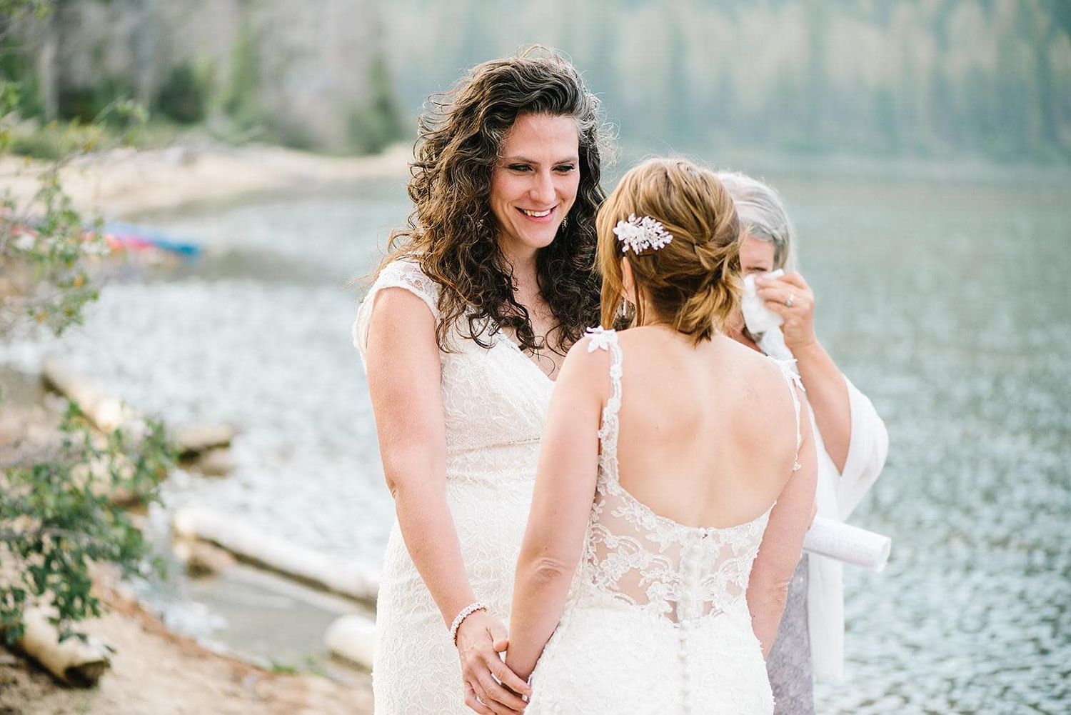 Two brides in white lace wedding dresses holding hands by a mountain lake on their wedding day.