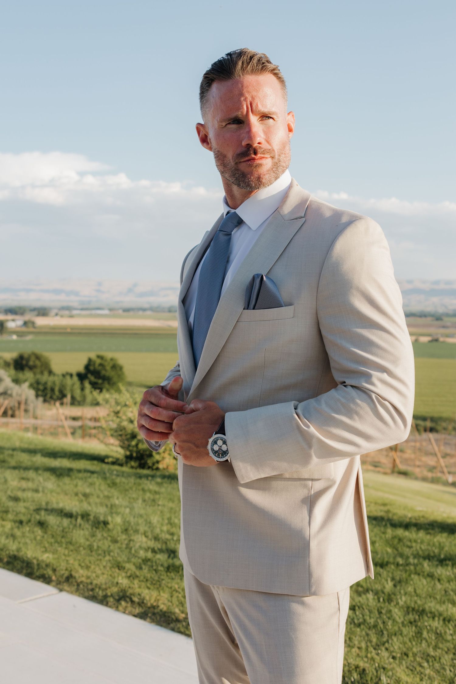 Handsome groom in tan suit with blue tie adjusting jacket outdoors with green fields in background.