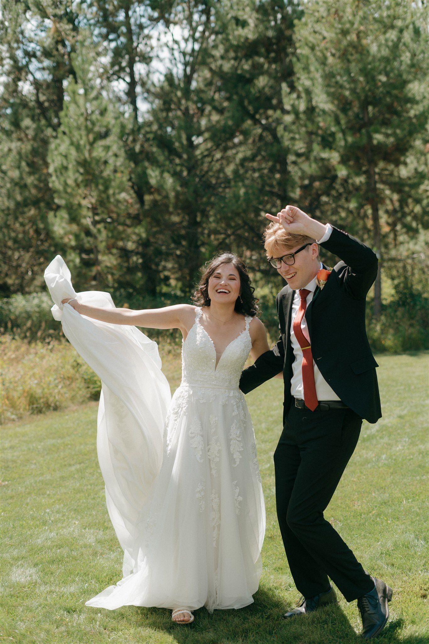 Joyful bride in lace gown twirling with laughing groom in black suit on green lawn surrounded by pine trees.