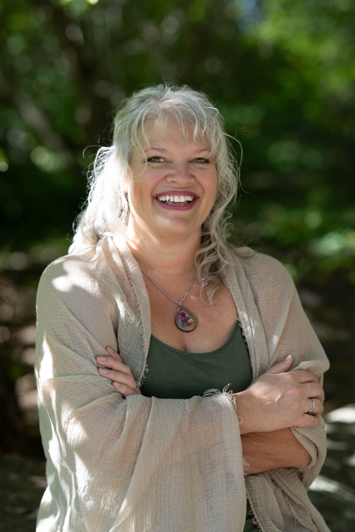 Smiling middle-aged woman with silver hair, arms crossed, wearing a beige cardigan and necklace outdoors.