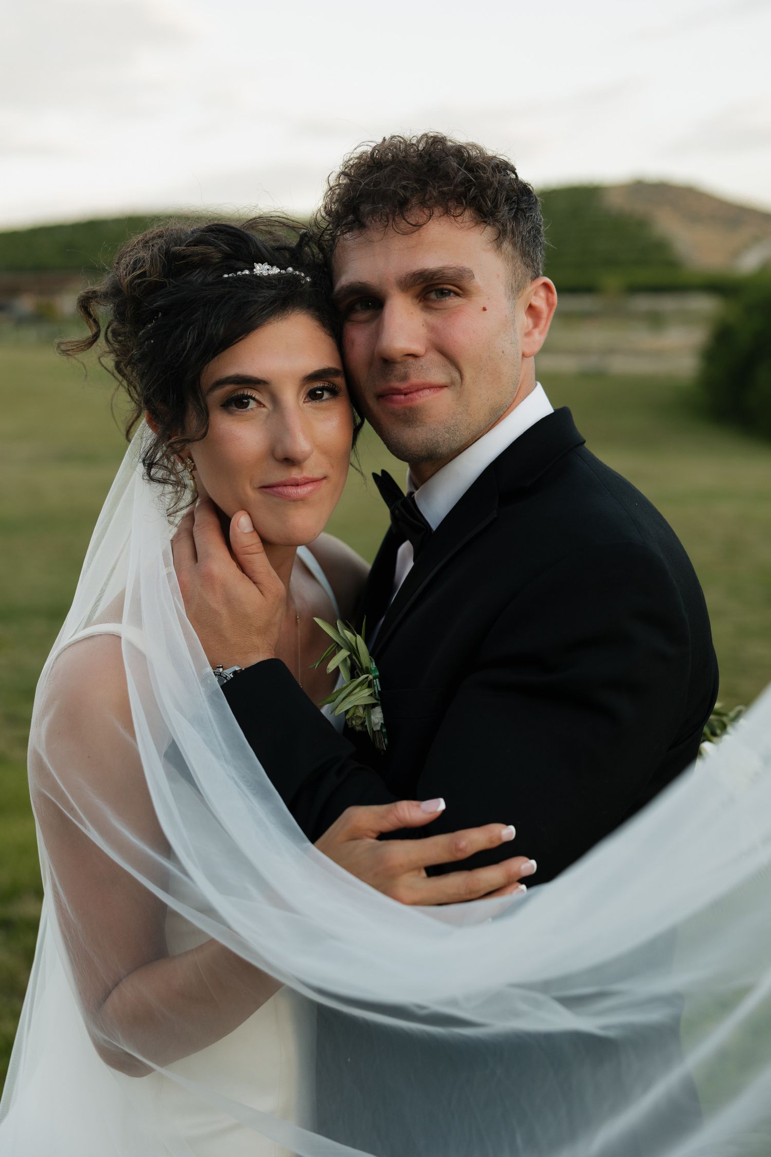 Newlywed couple embraces outdoors, bride in veil leans against groom in black tuxedo with green hills behind.