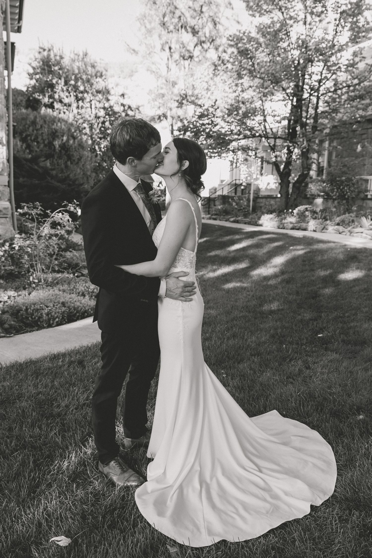 Black and white photo of bride and groom kissing in garden wearing elegant wedding attire.