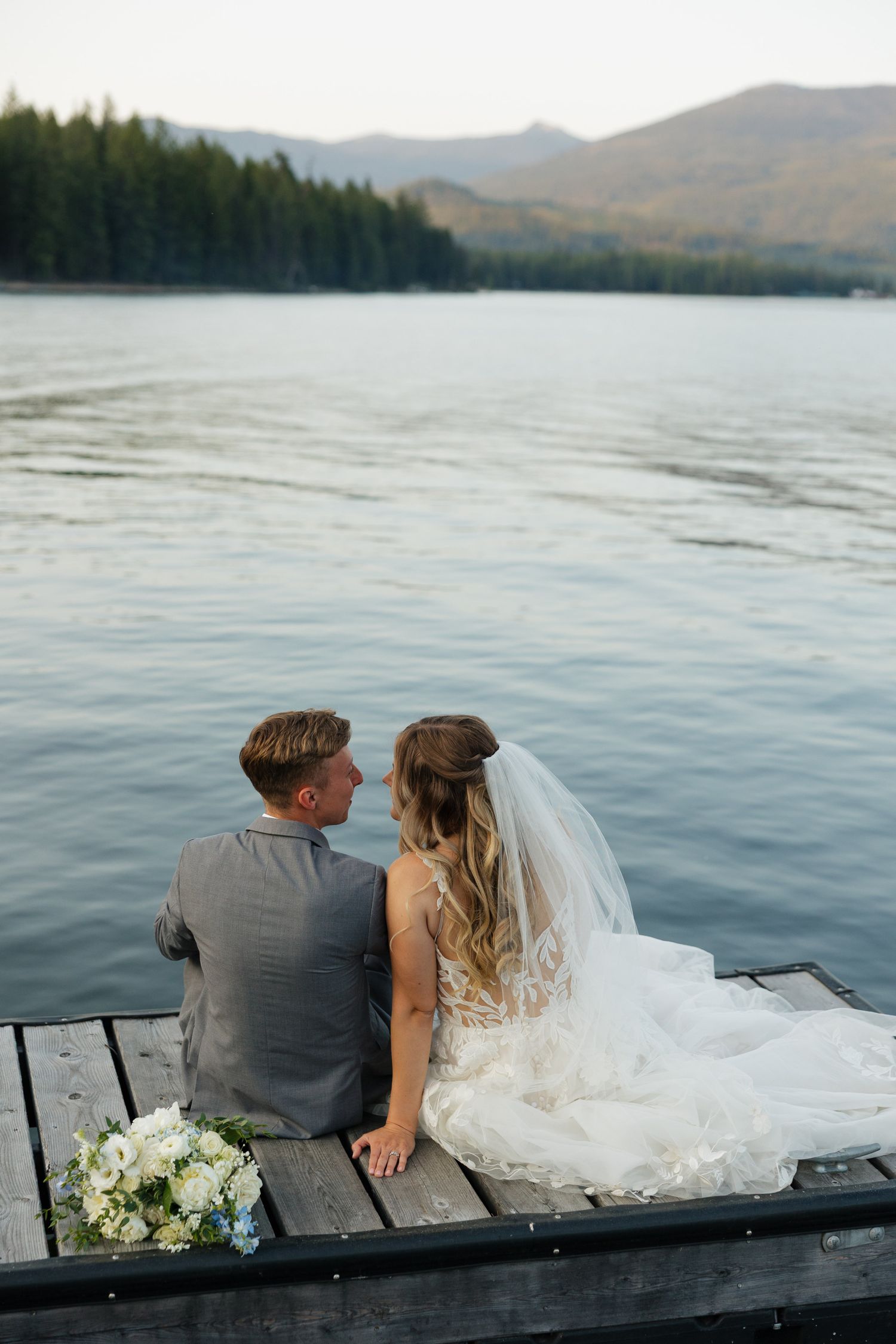 Bride and groom sit on wooden dock holding hands, gazing at serene mountain lake at sunset.