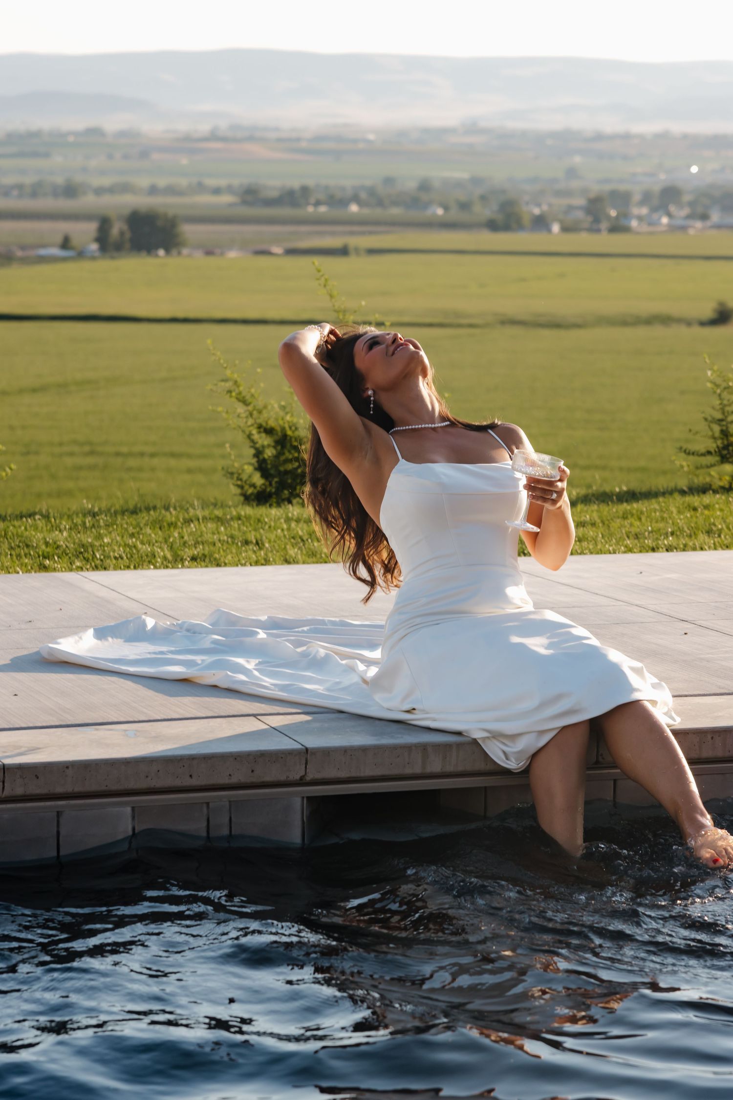 Bride in white dress sitting poolside with feet in water, scenic green countryside landscape behind her.