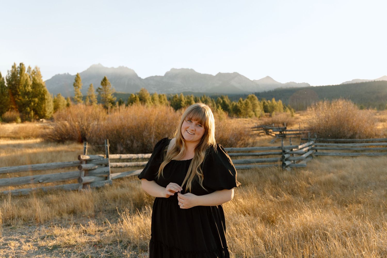 Smiling blonde woman in black dress poses in golden field with mountain range backdrop at sunset.