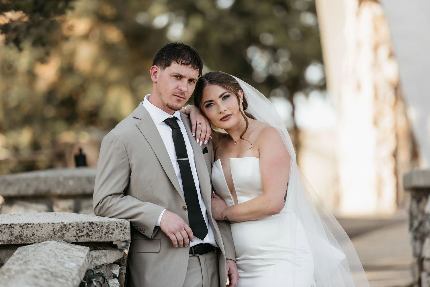 Bride in white gown and groom in beige suit posing together against stone wall in soft golden outdoor light.