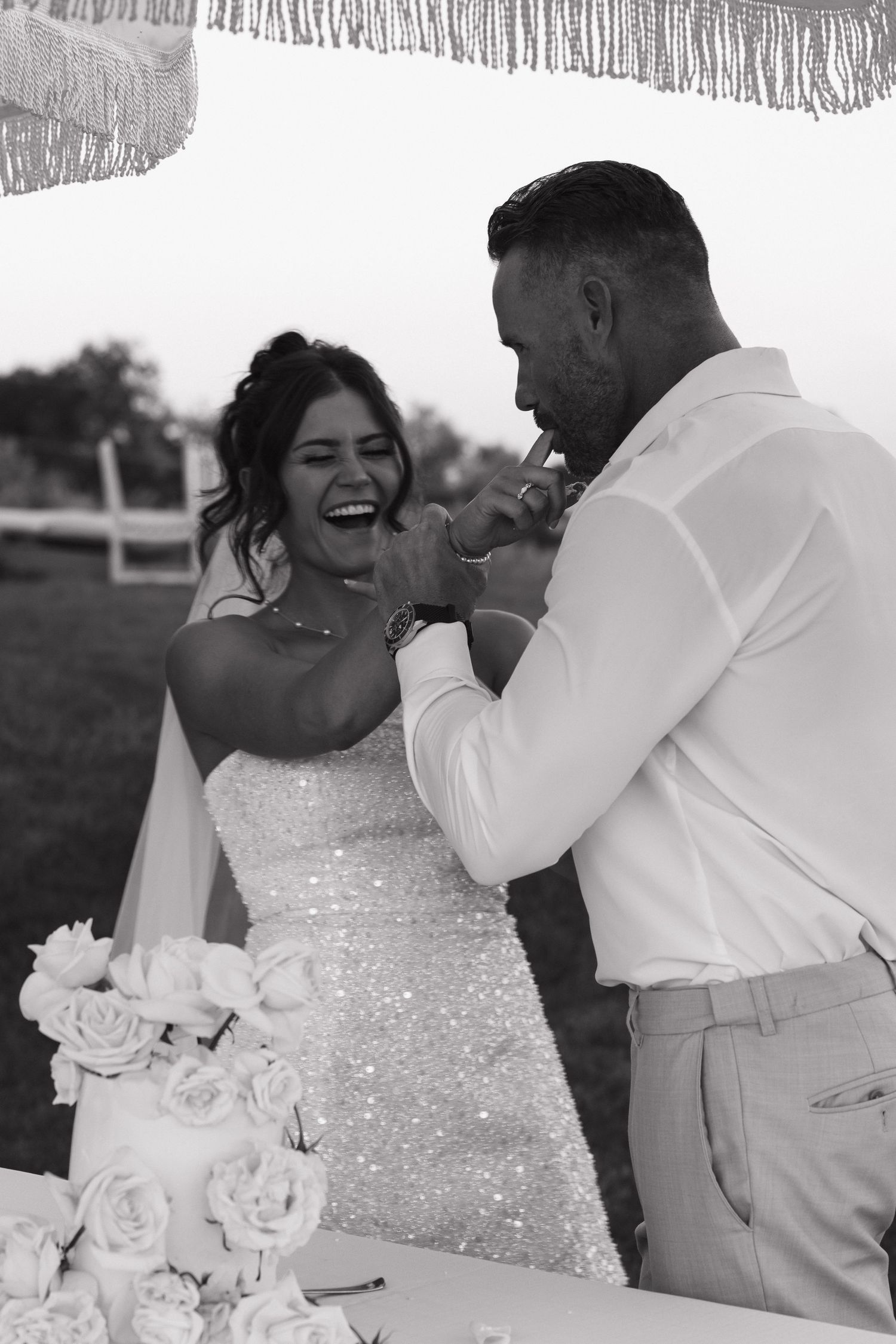 Black and white photo of laughing bride and groom cutting their wedding cake outdoors at sunset.