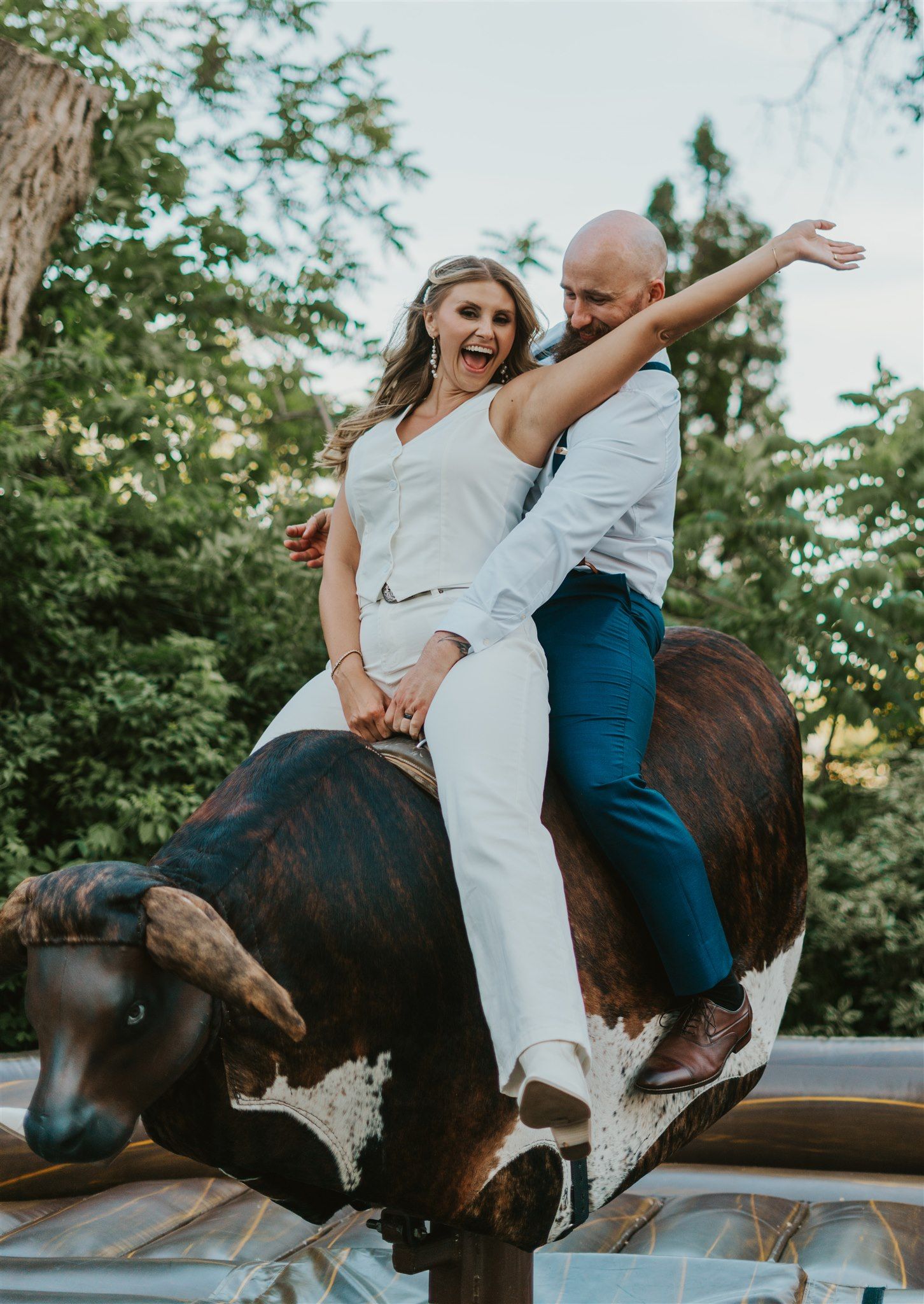 Excited couple riding mechanical bull outdoors, bride in white jumpsuit laughing with arms raised in celebration.