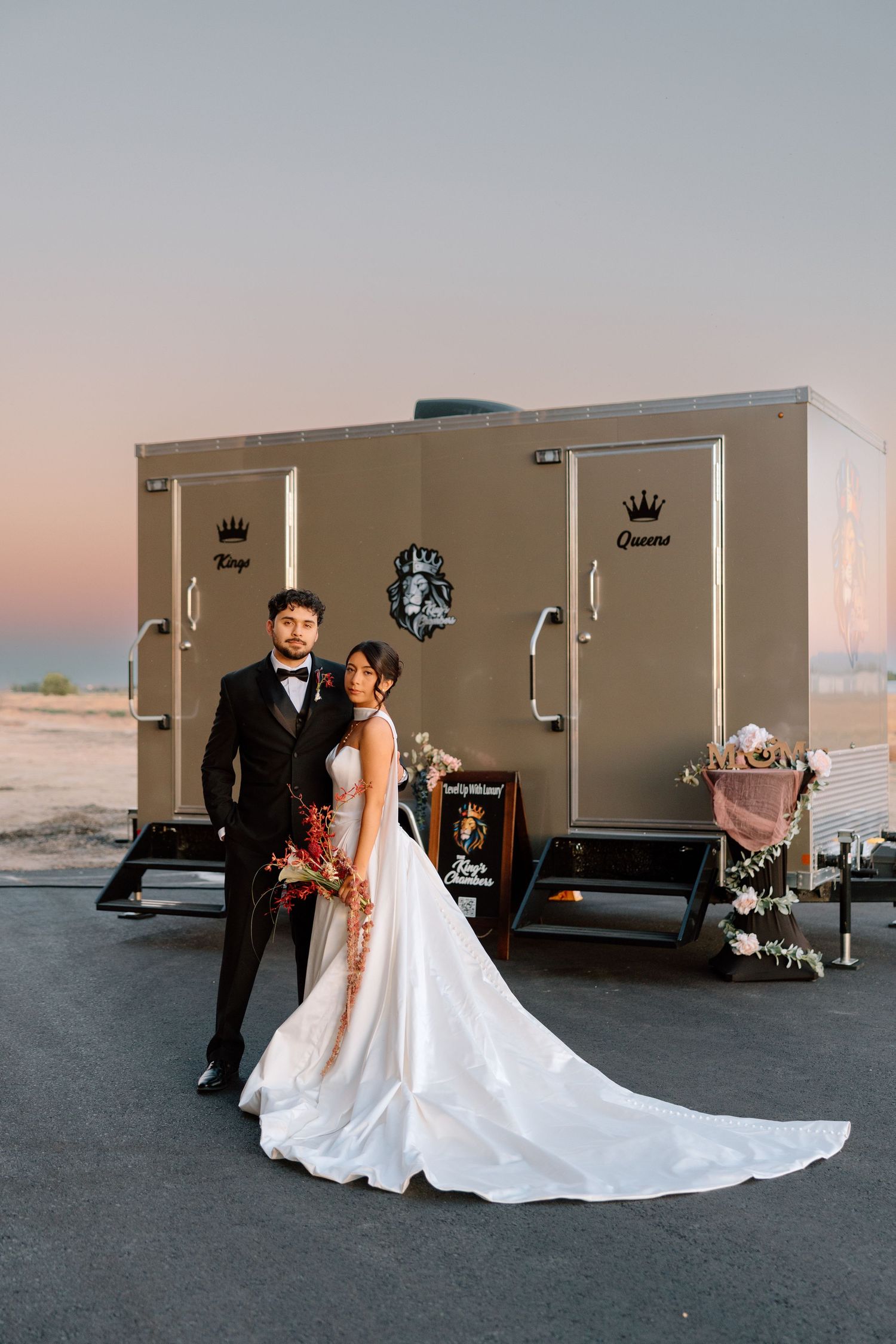 Bride and groom pose in formal wedding attire before a luxury portable restroom trailer at sunset.