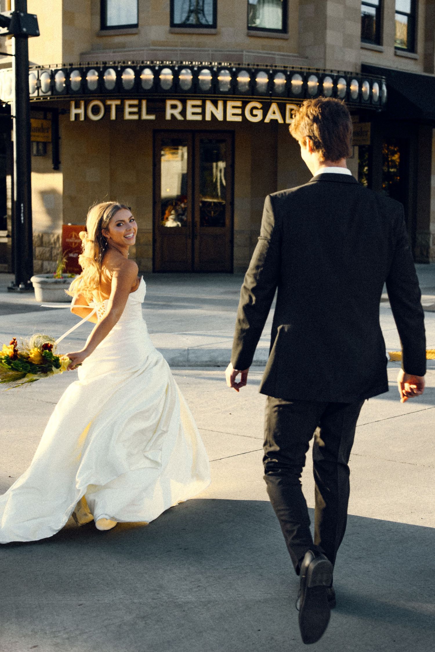 Bride in white gown smiles back at groom walking toward Hotel Renegade entrance at sunset.