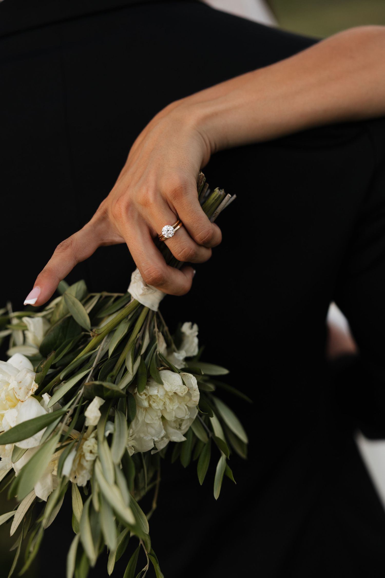 Bride wearing diamond solitaire ring holds white peony and greenery bouquet against black fabric background.