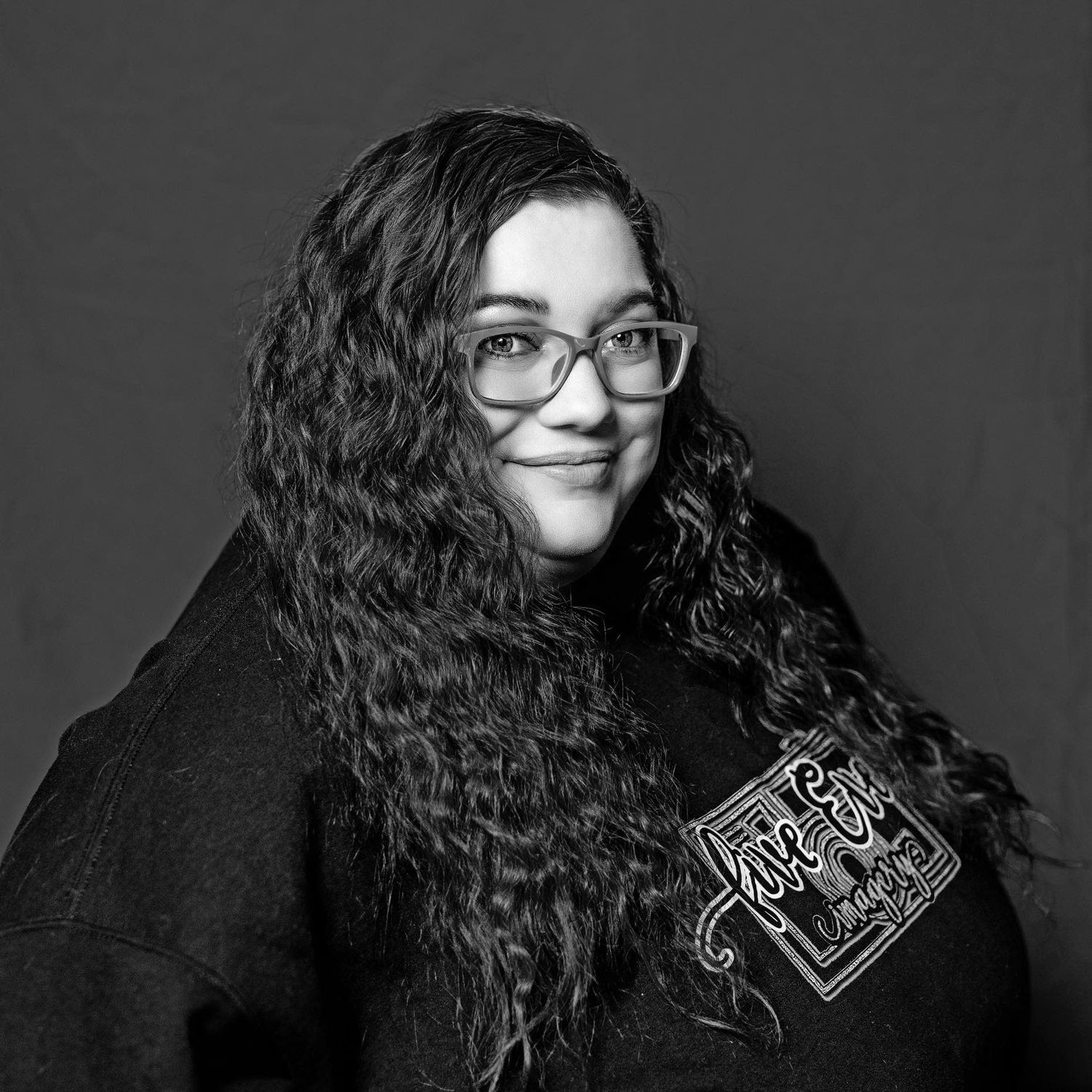 Black and white portrait of a curly-haired woman wearing glasses and dark clothing against gray backdrop.