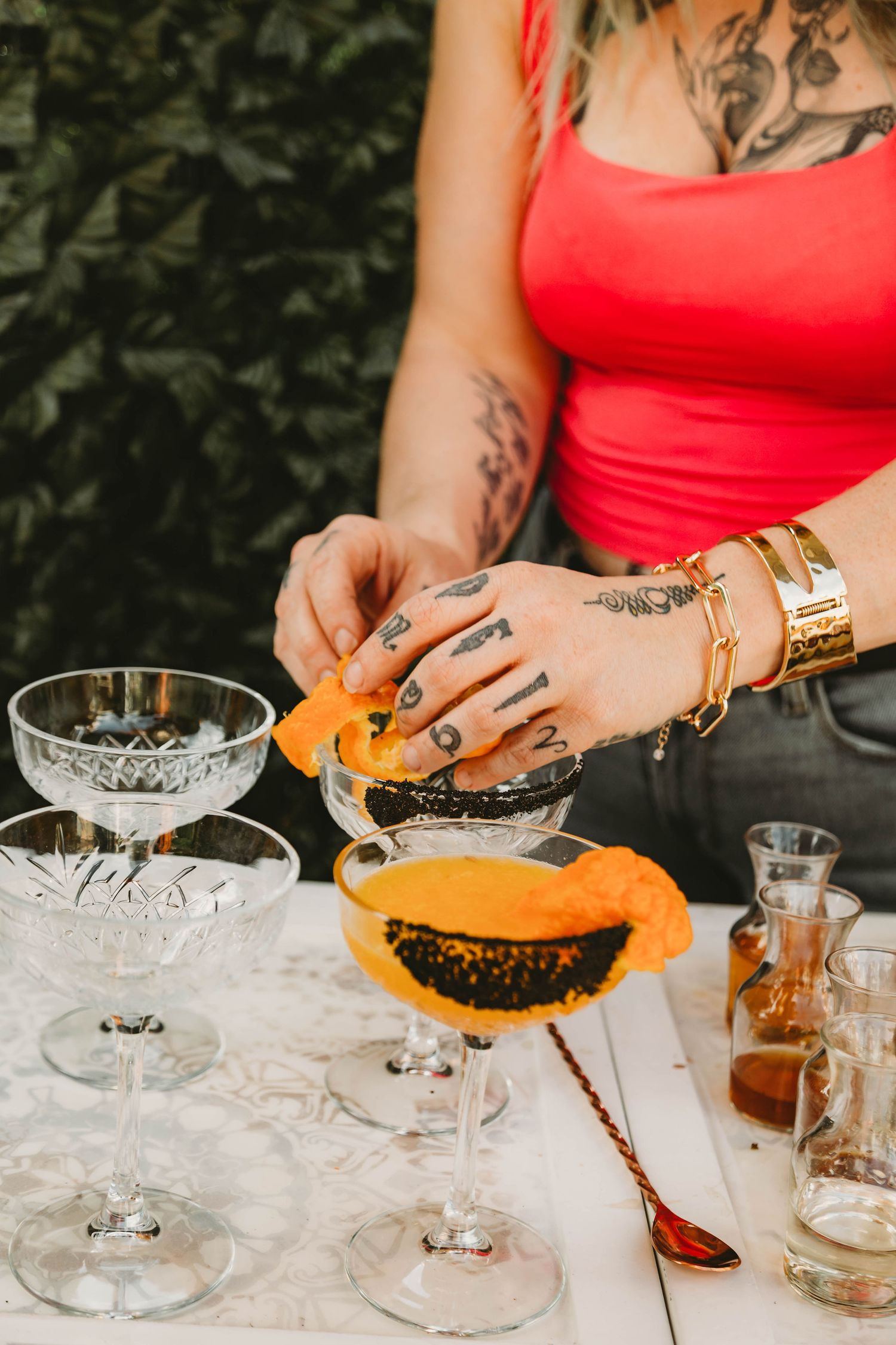 Tattooed bartender garnishing cocktails with orange peel slices in crystal coupe glasses outdoors.