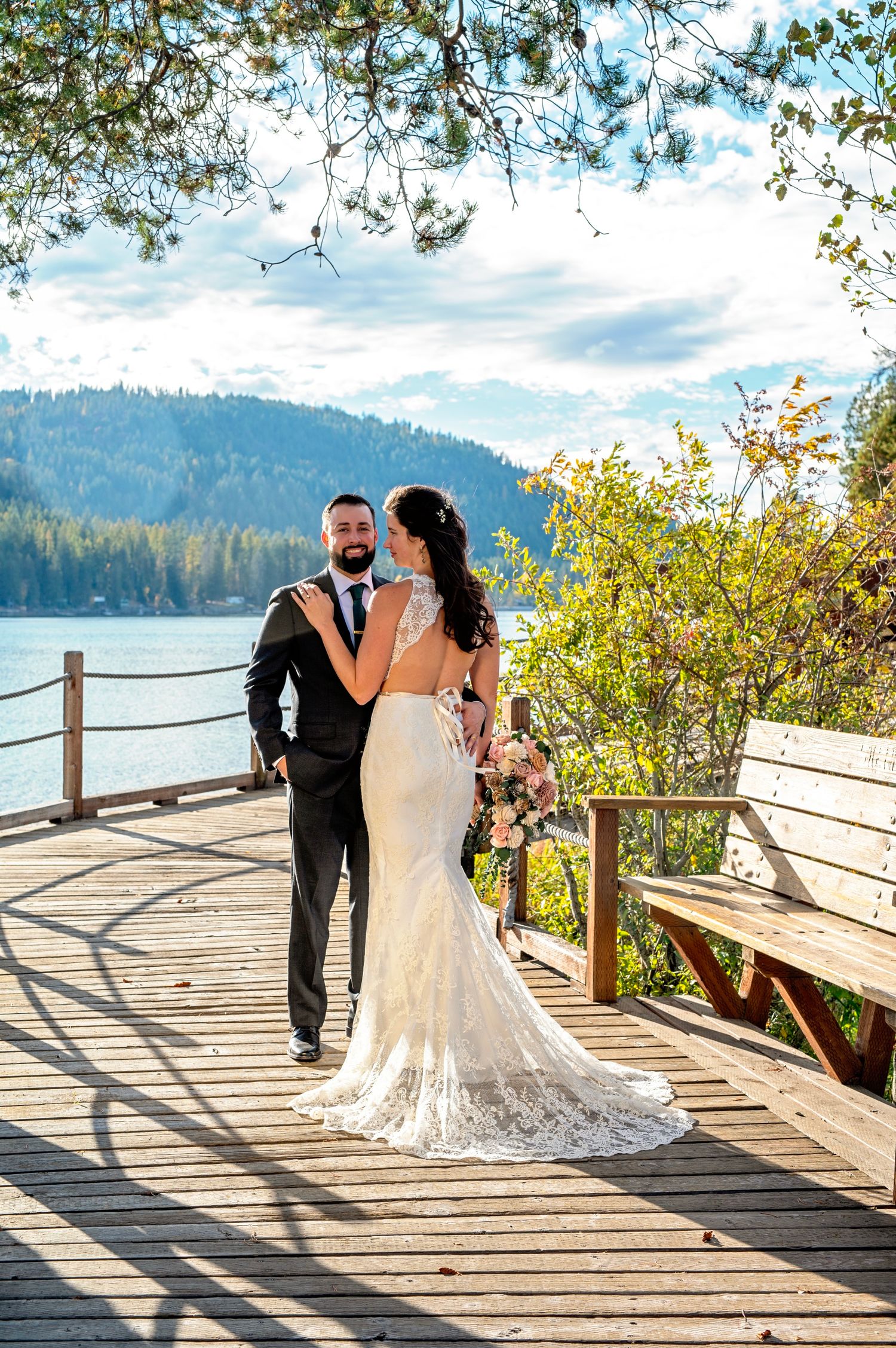 Bride in lace gown and groom in black suit pose on a lakeside dock surrounded by autumn trees.