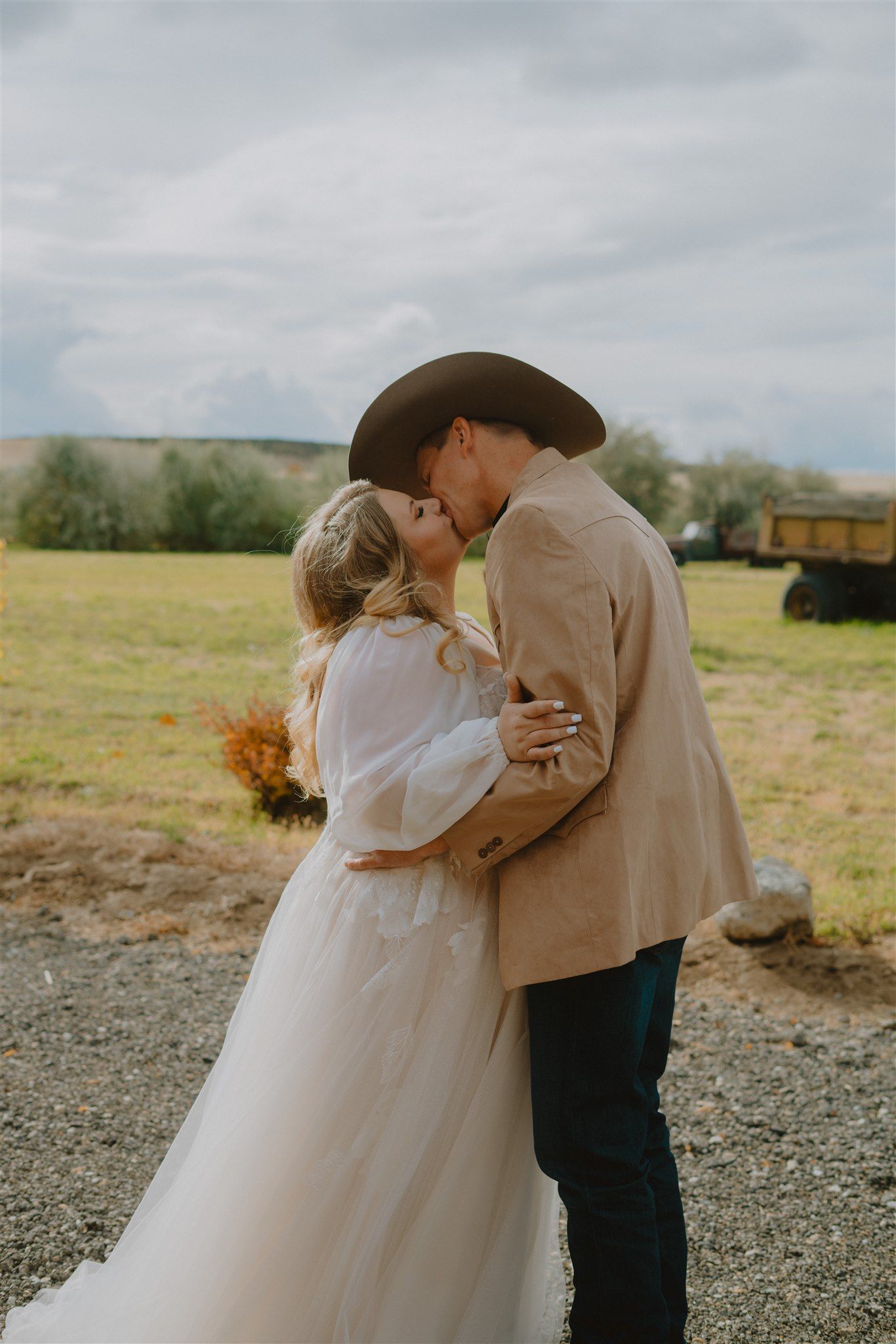 Bride and groom sharing a romantic kiss outdoors, groom wearing cowboy hat and tan jacket, bride in flowing white gown.