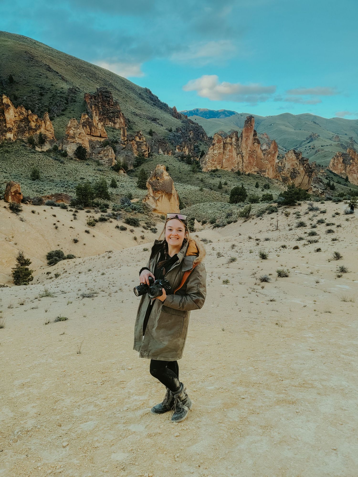 Woman smiling in olive trench coat holding camera with dramatic rocky desert landscape and mountains behind her.