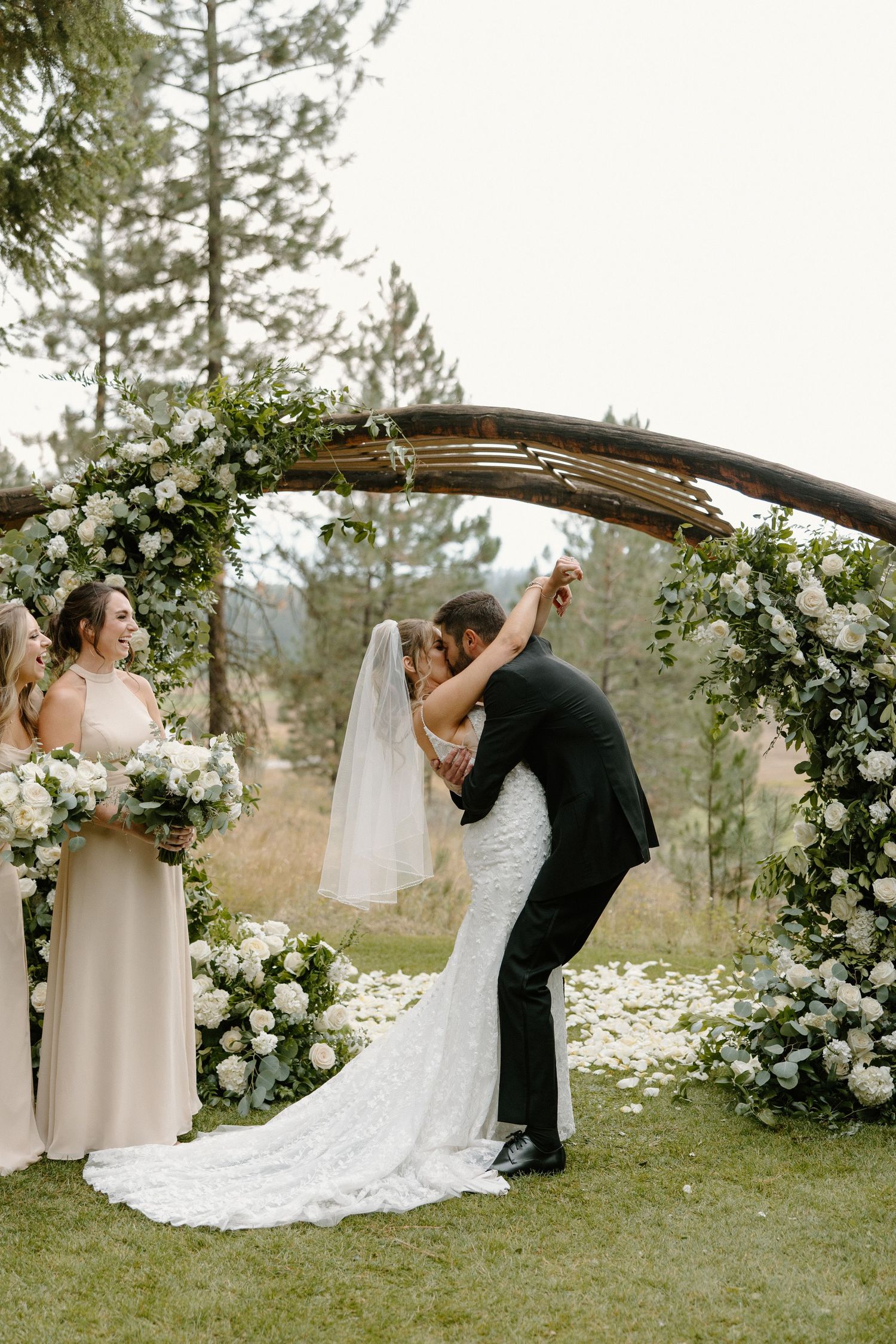 Bride and groom share first kiss under a rustic wooden arch decorated with white flowers and greenery outdoors.