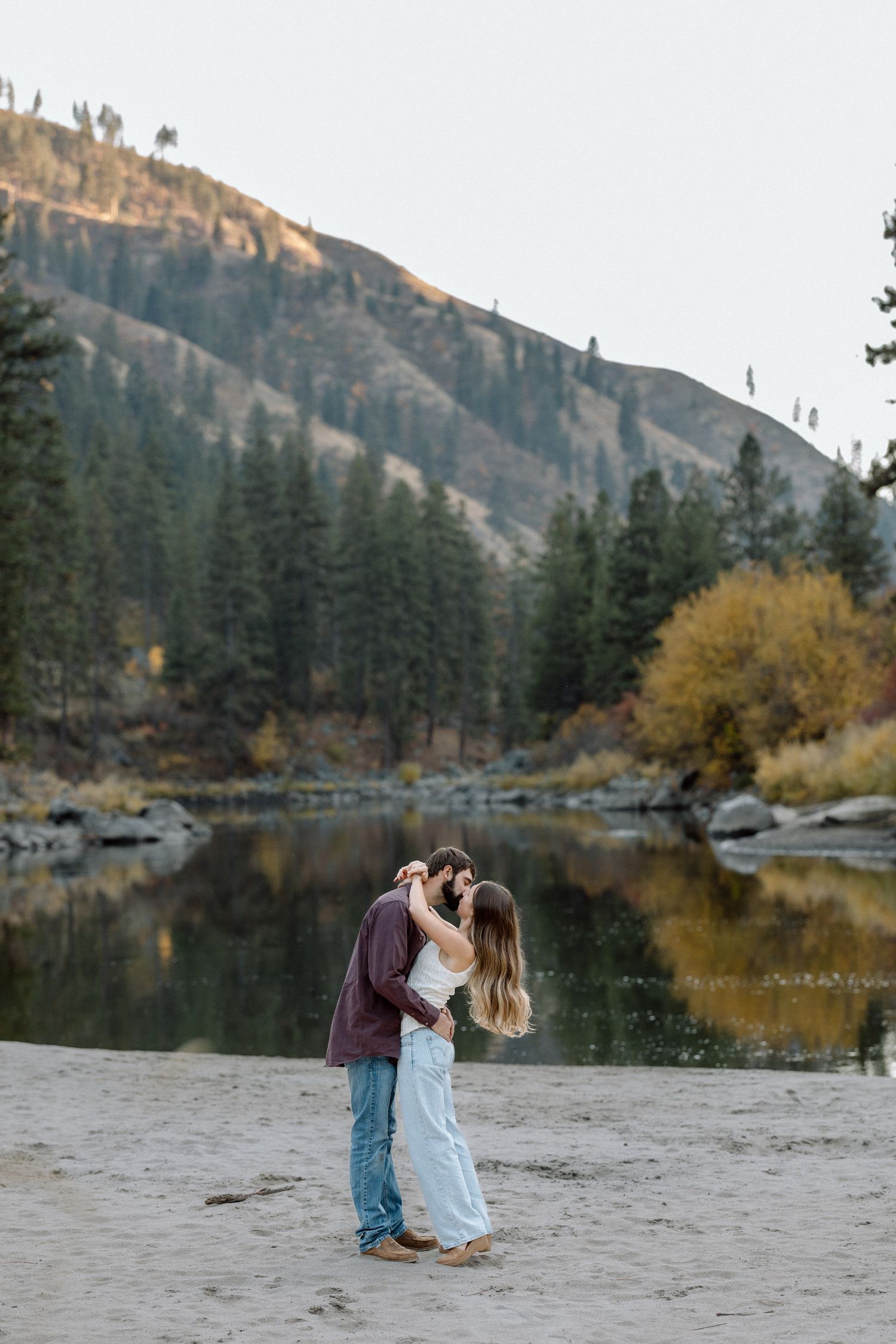 Couple sharing a romantic kiss on a sandy riverbank with mountain reflections and autumn foliage backdrop.