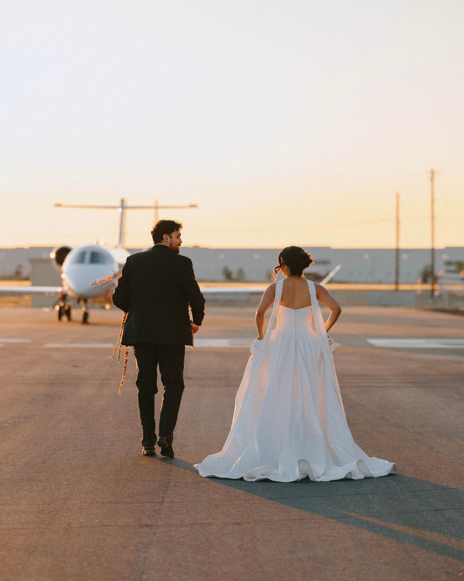 Bride and groom walking toward a private jet on a runway at sunset, bride in elegant white gown with train.