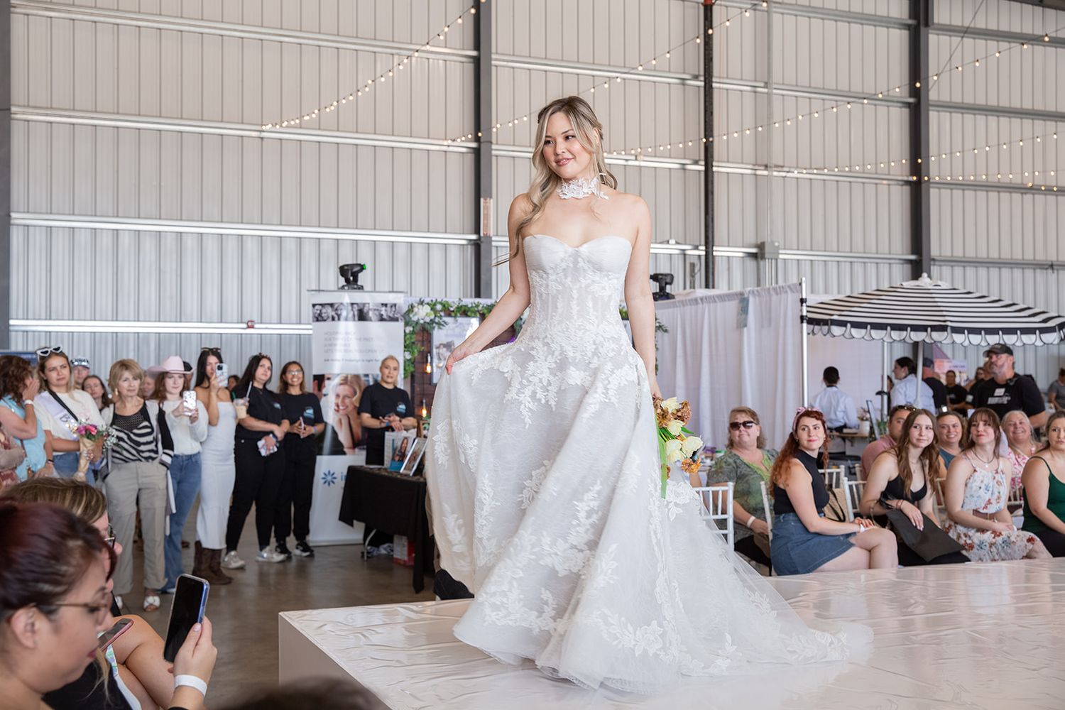 Bride modeling a strapless lace ballgown wedding dress on a runway at an indoor bridal fashion show.