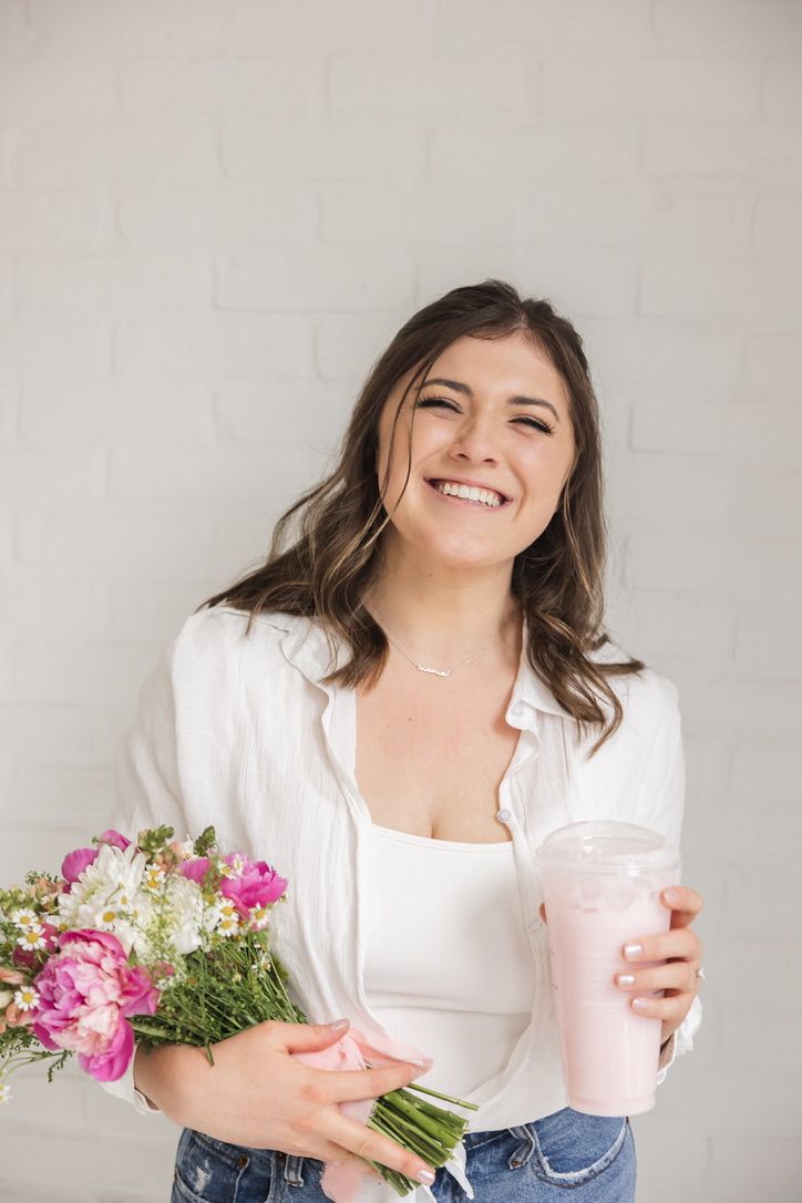 Smiling brunette woman in white shirt holding pink flowers bouquet and pink smoothie against white brick wall.