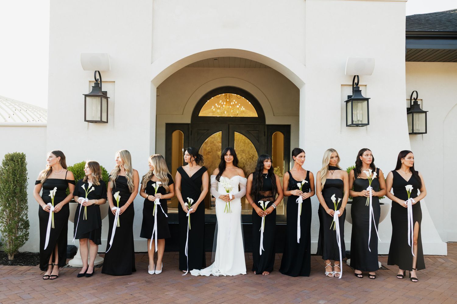Bride in white gown poses with bridesmaids in black dresses outside elegant white venue with arched entrance.