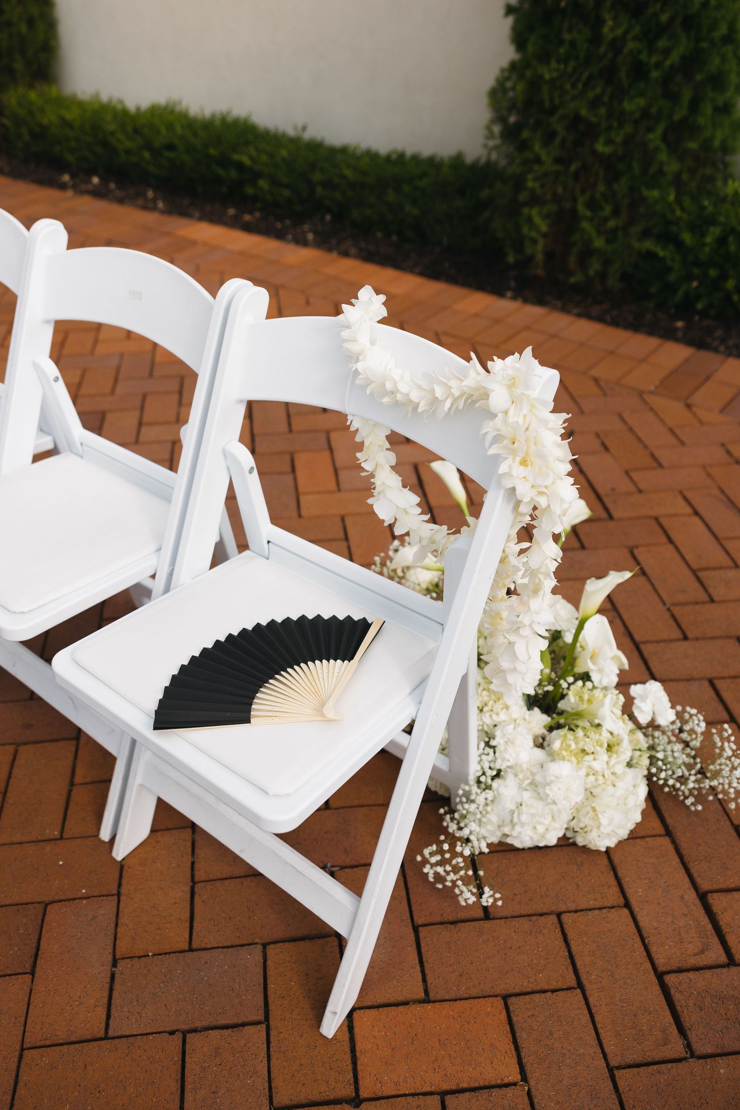 White folding chairs with black fan and white floral arrangement on brick pathway.
