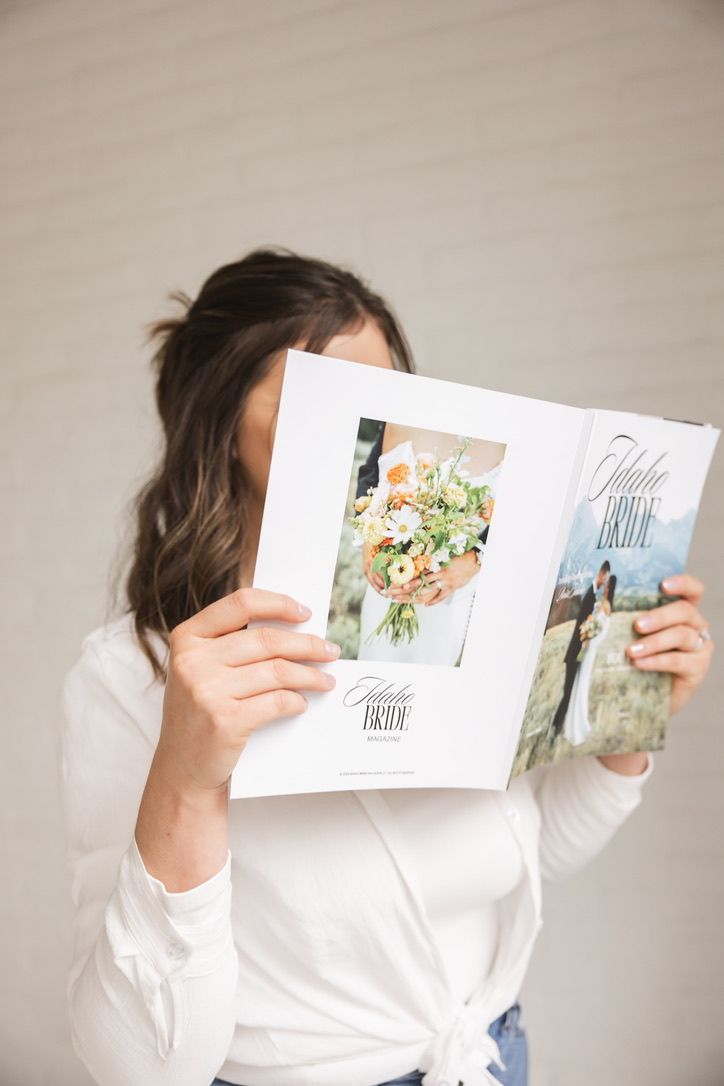Woman holding Idaho Bride magazine open, covering her face, wearing white top and jeans.