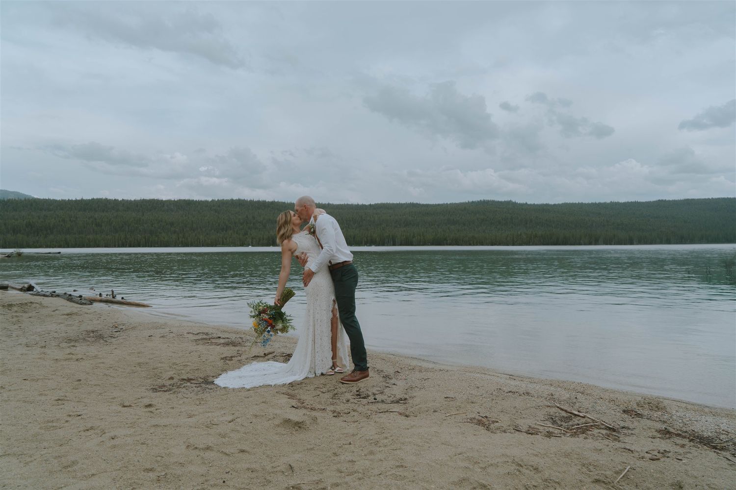 Bride and groom kissing on sandy lakeshore with serene water and green hills in the background on cloudy day.