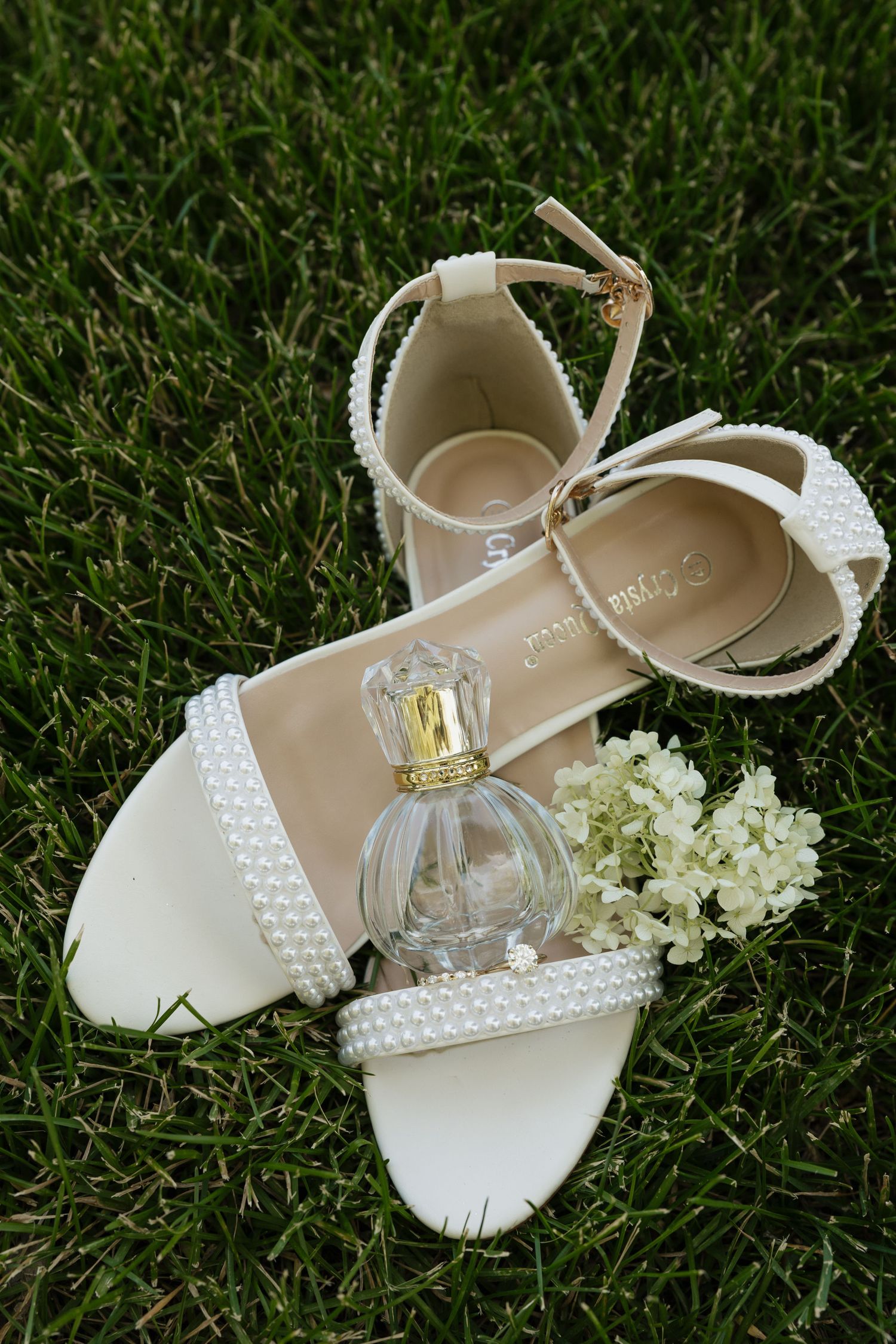 White pearl wedding sandals with ankle strap beside perfume bottle and white flowers on green grass.