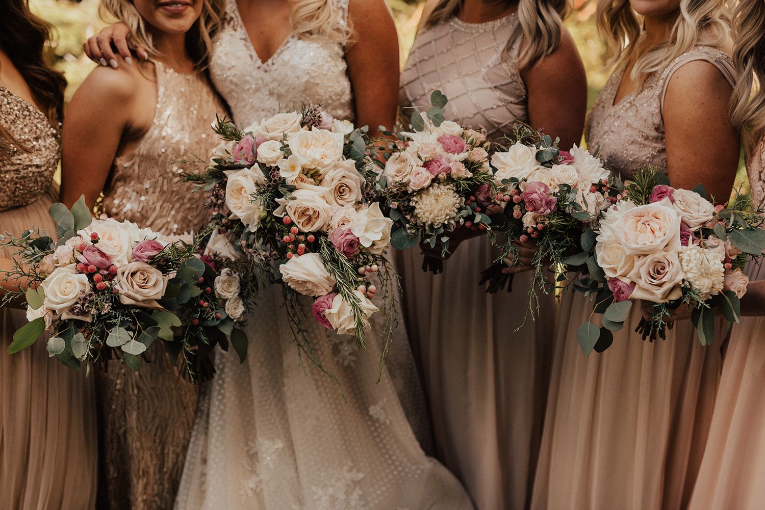 Bride and bridesmaids holding pink and white rose bouquets in taupe gowns.