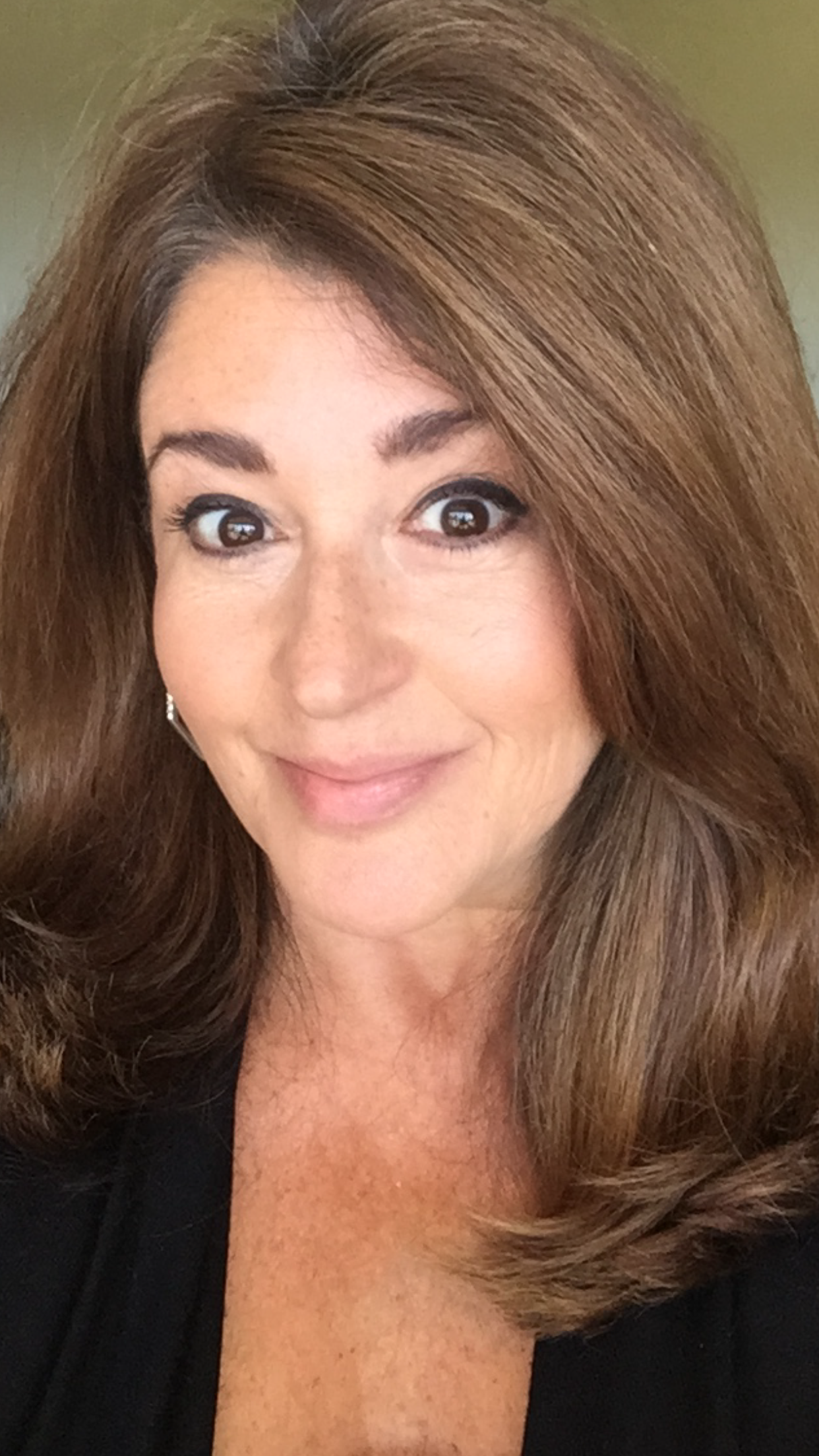 Smiling woman with brown shoulder-length hair taking a selfie wearing a black top in a warmly lit setting.