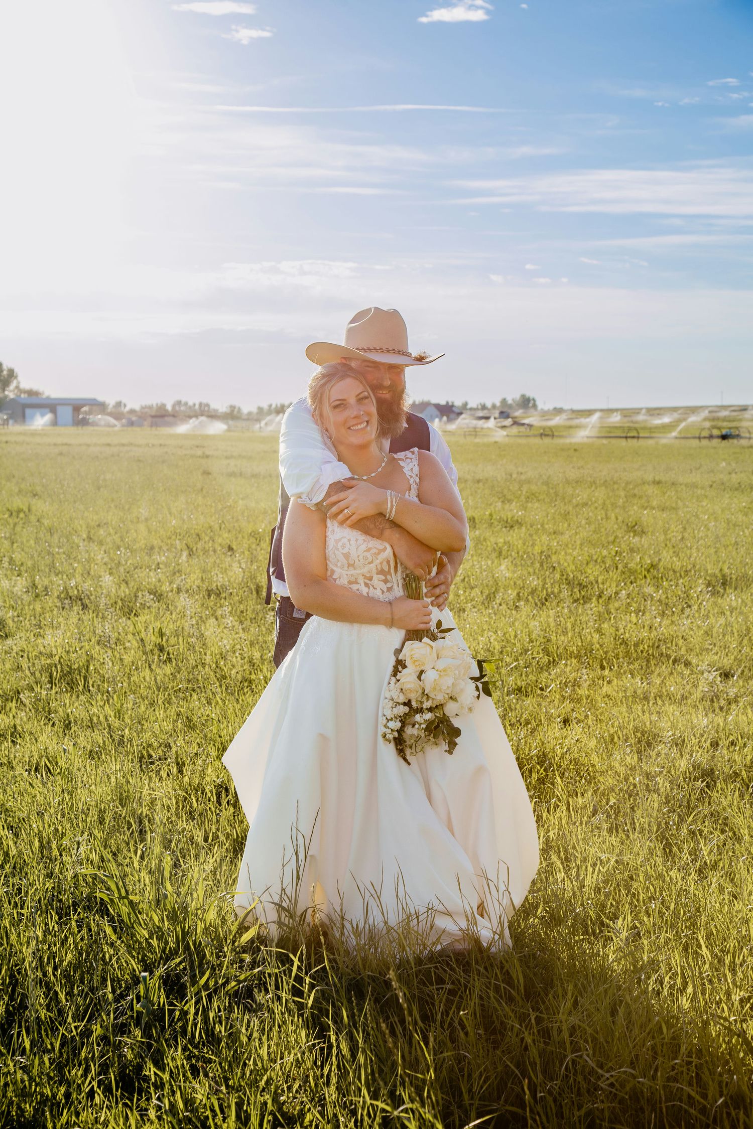 Bride and groom embracing in a sunny green field, groom wearing a cowboy hat, bride holding a white bouquet.