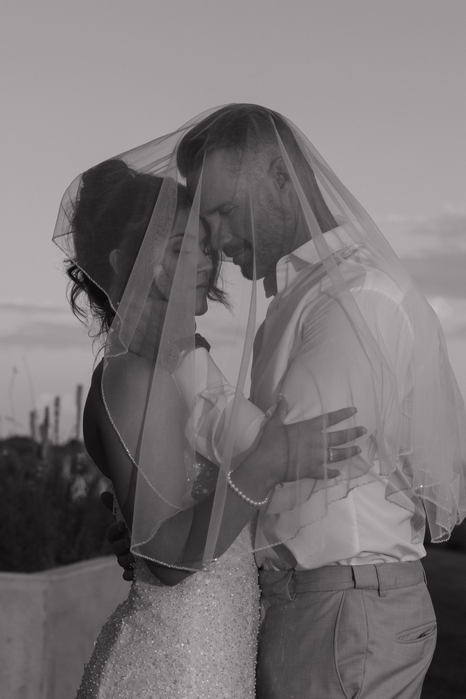 Romantic black and white photo of bride and groom sharing intimate moment under wedding veil at dusk.