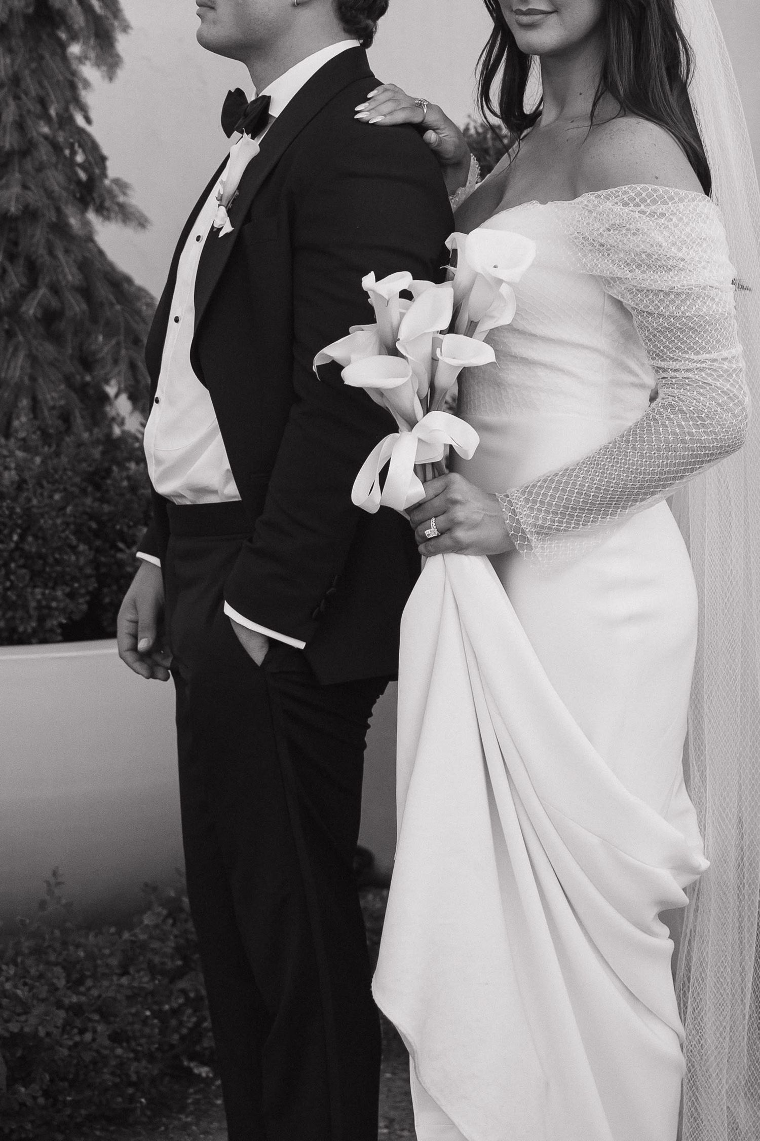 Black and white photo of bride in off-shoulder gown holding calla lily bouquet beside groom in tuxedo.