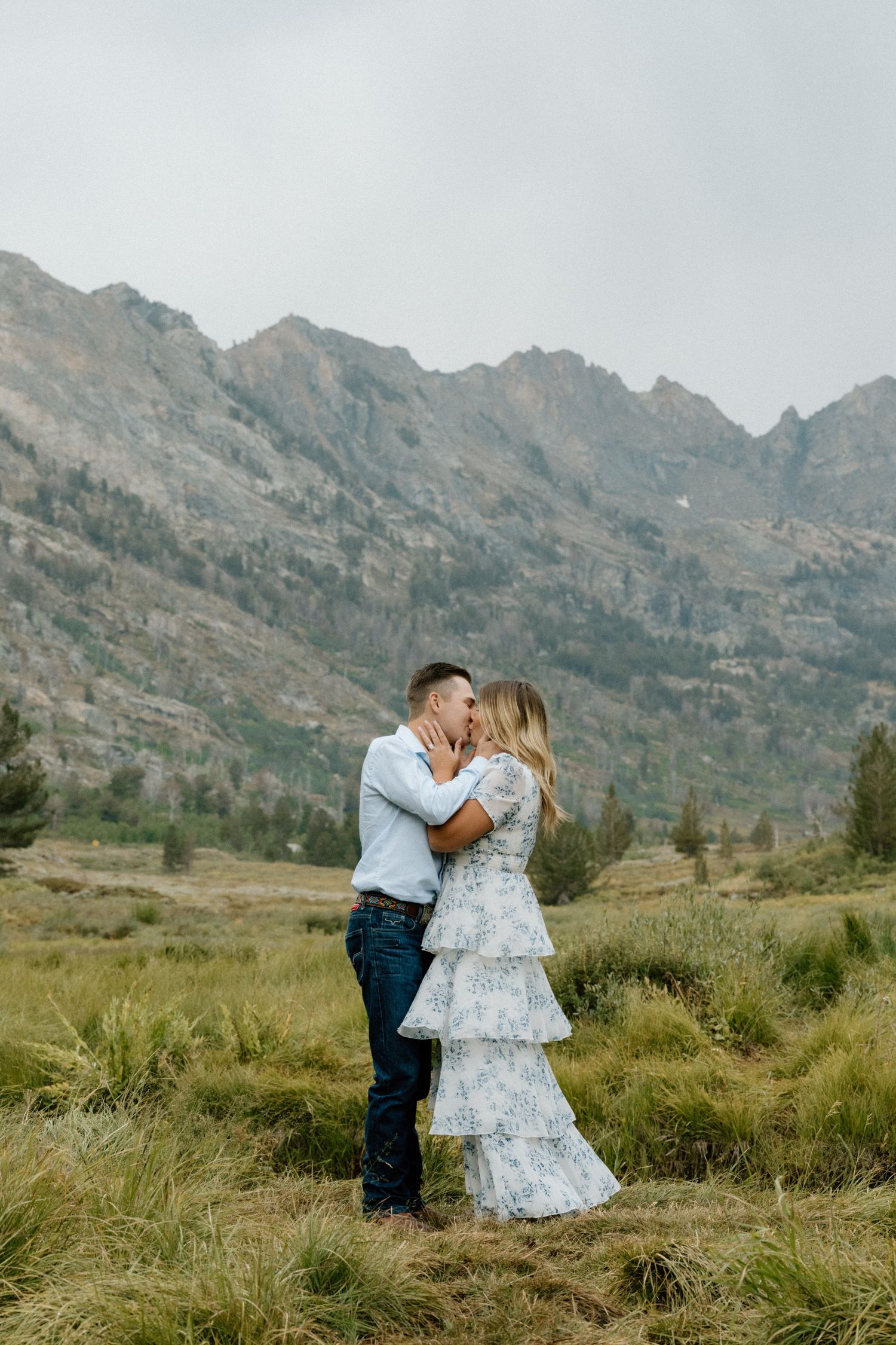 Couple embracing and kissing in a mountain meadow with dramatic rocky peaks and overcast skies behind them.