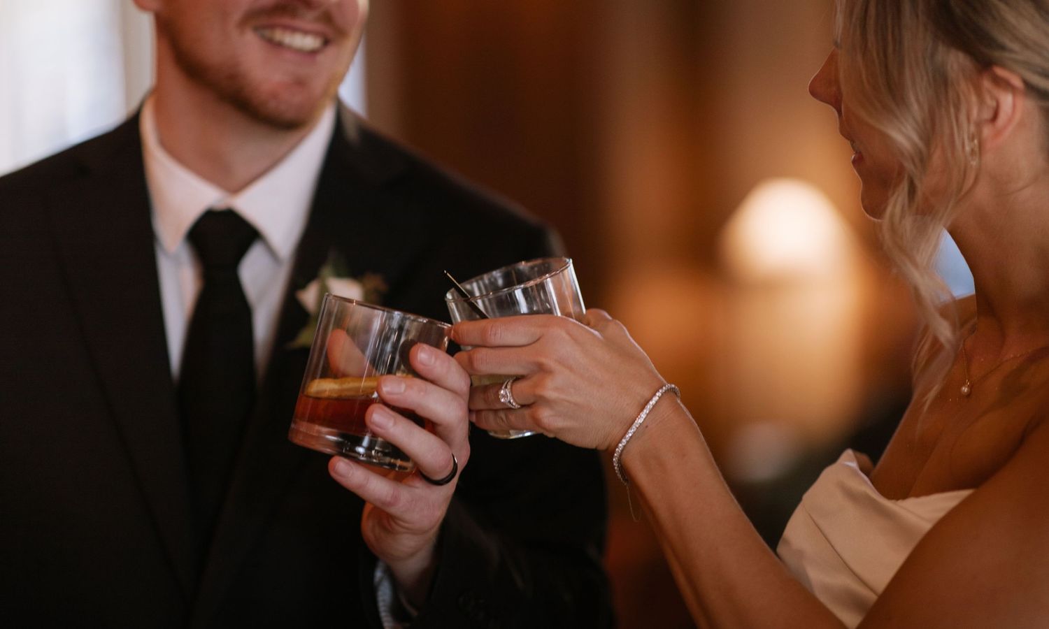 Bride and groom clinking whiskey glasses in a warm romantic setting.