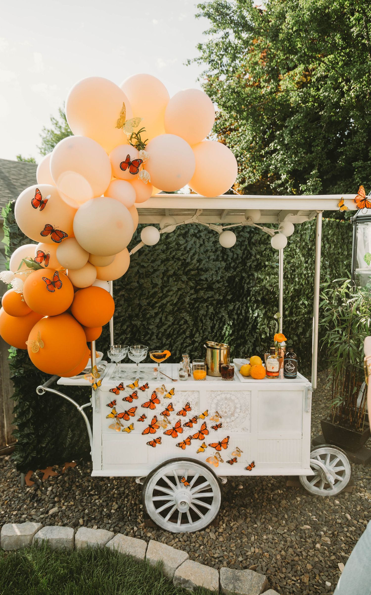 White vintage cart decorated with orange and peach balloon garland and monarch butterfly decorations for an outdoor party.