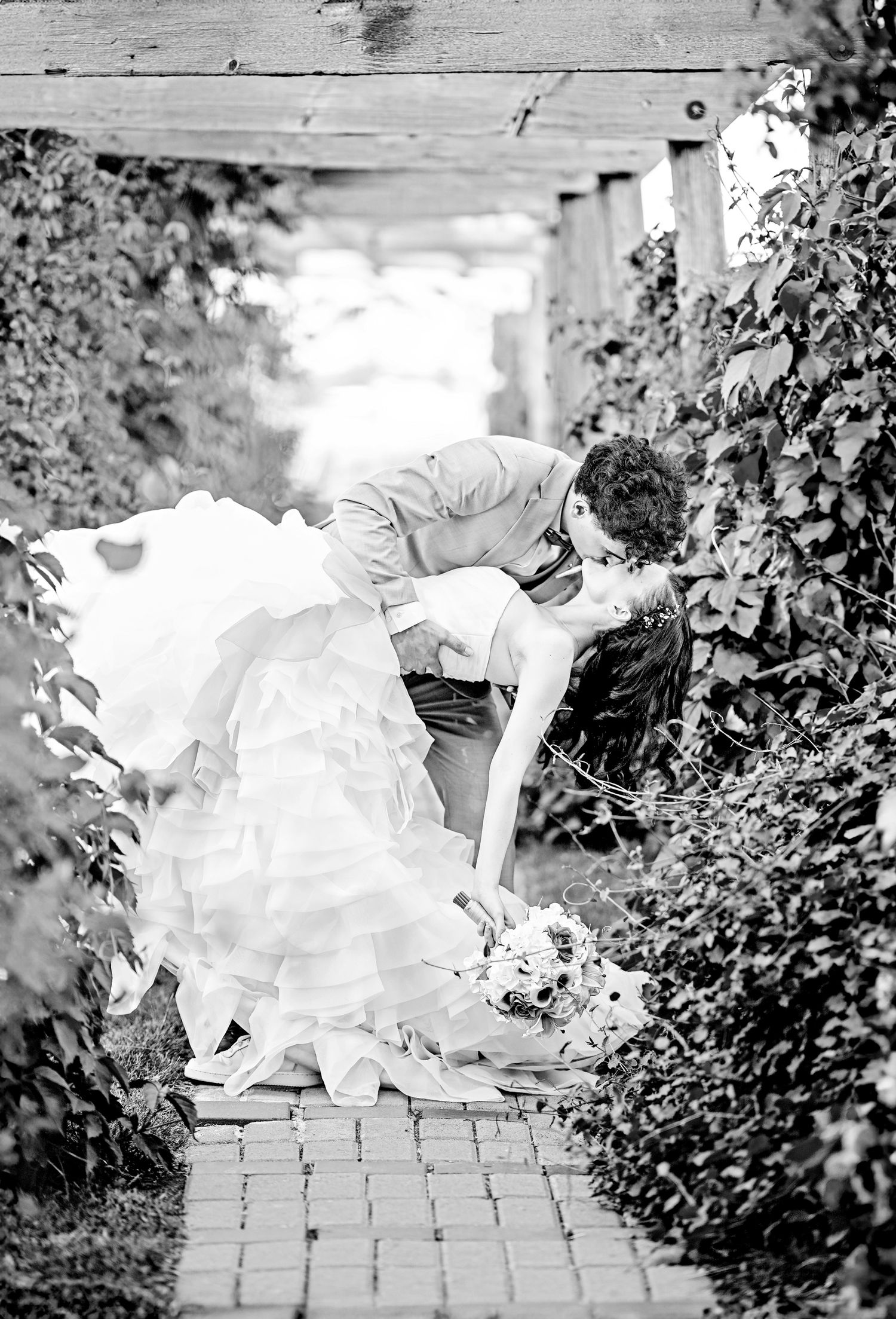 Black and white photo of groom dipping bride for a kiss on a brick path surrounded by ivy.