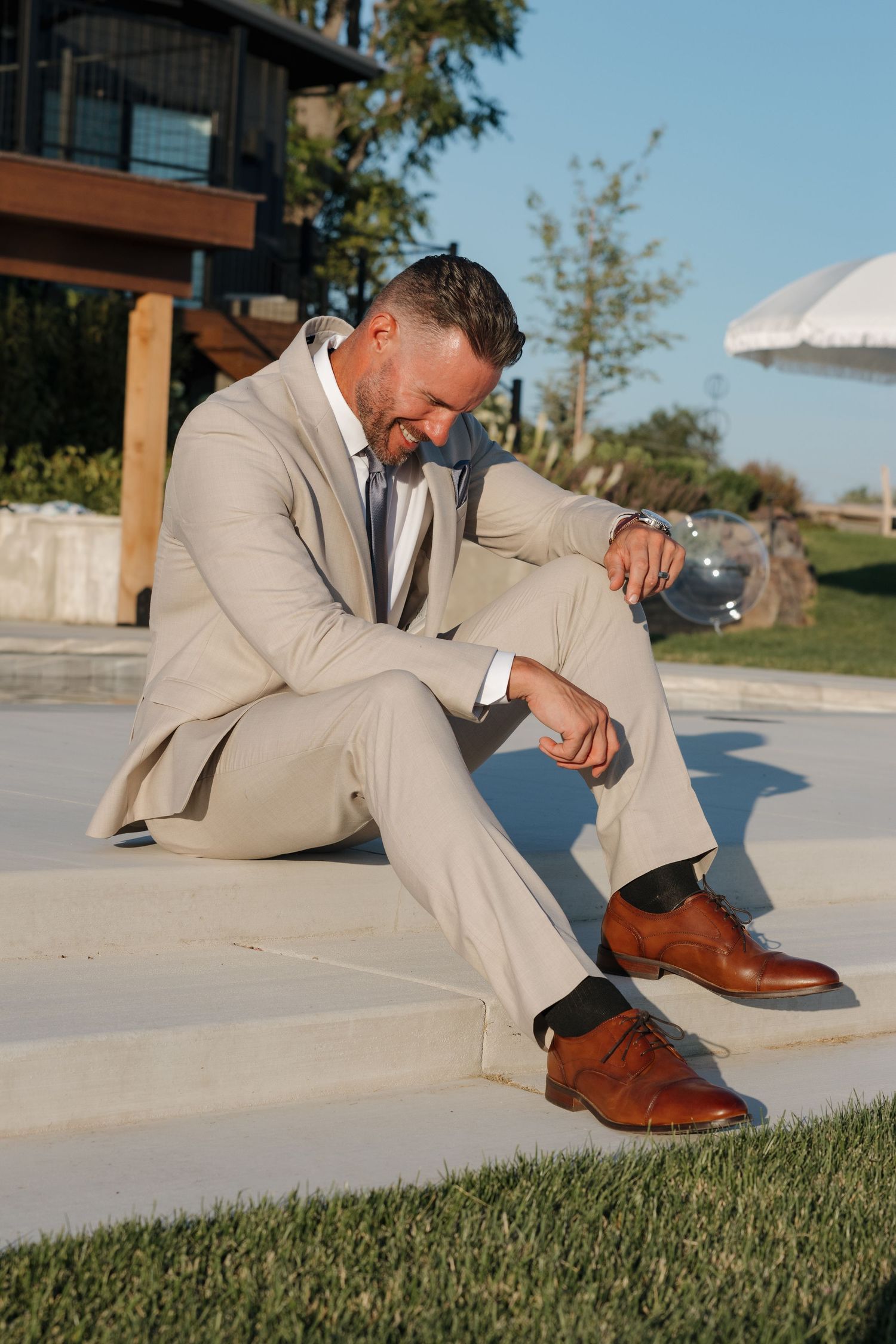 Smiling groom in beige suit sitting outdoors by pool holding champagne glass at wedding celebration.